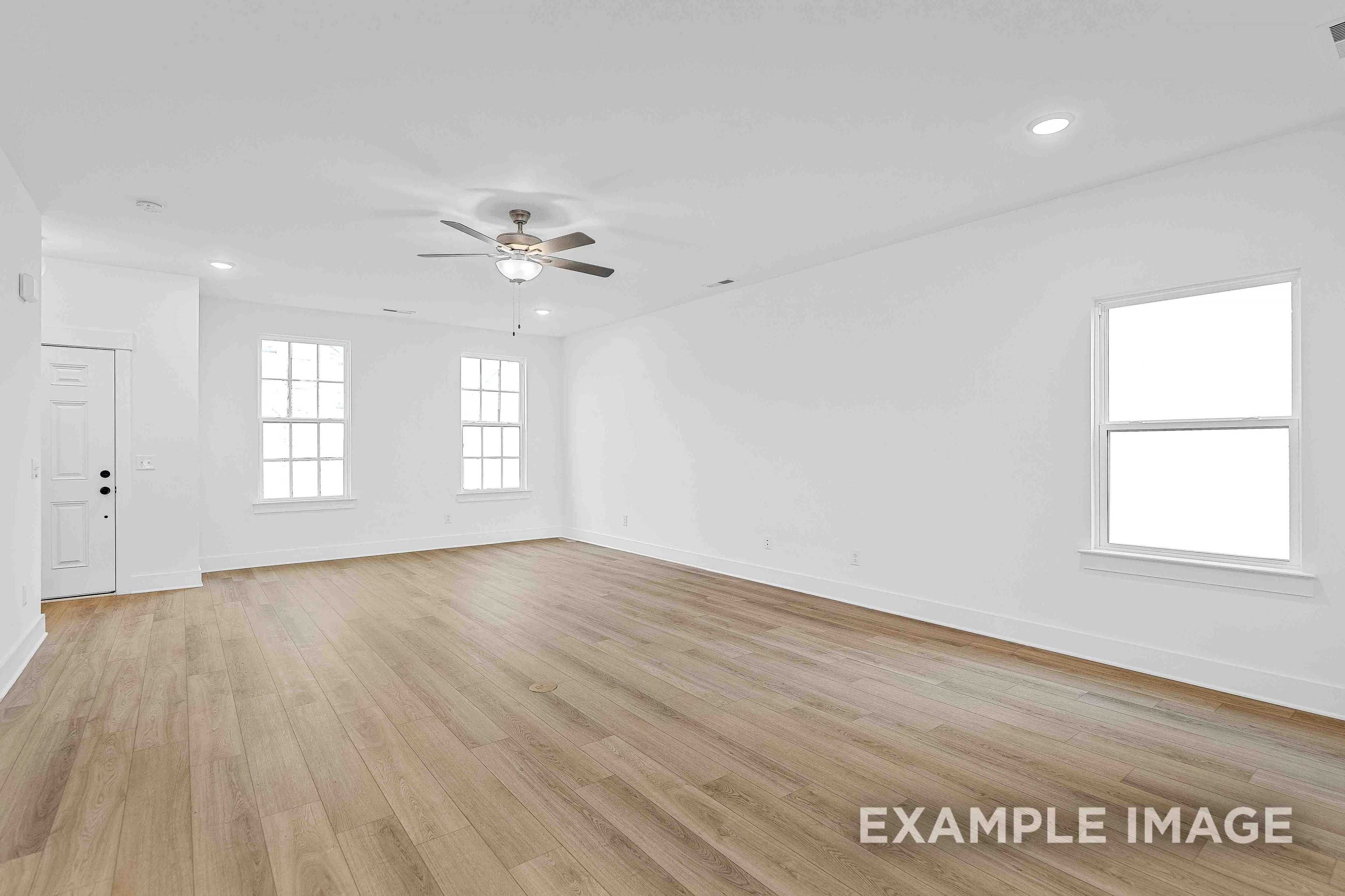 Spacious empty living room in The Charleston C home design with light hardwood floors, white walls, ceiling fan, and large windows