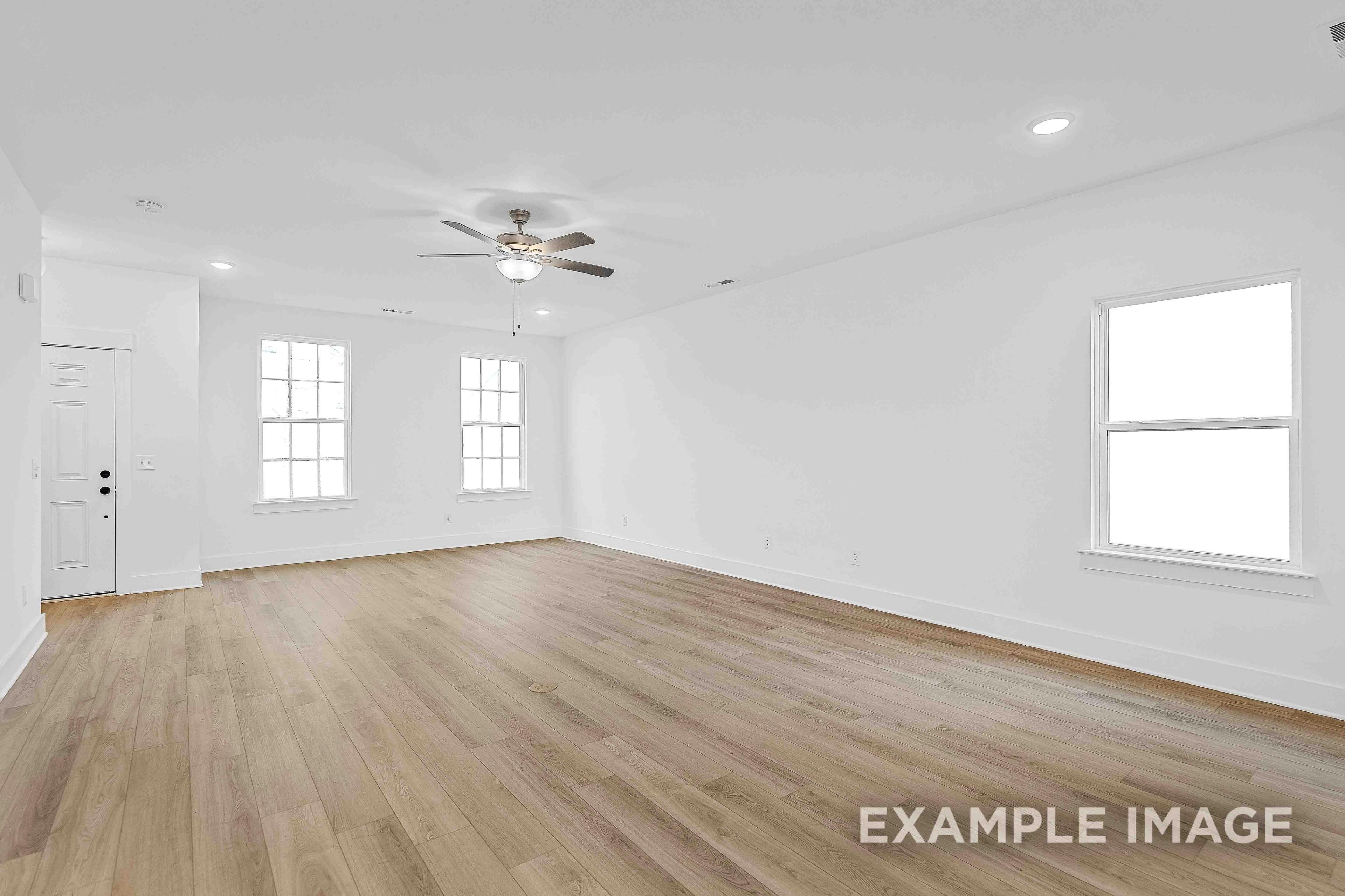 Spacious empty living room in The Charleston C home design with light hardwood floors, white walls, ceiling fan, and large windows