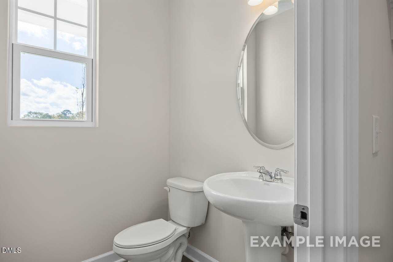 Bright half bath with round mirror, white pedestal sink, toilet, and window in Davidson Homes The Graham, Fuquay-Varina, NC