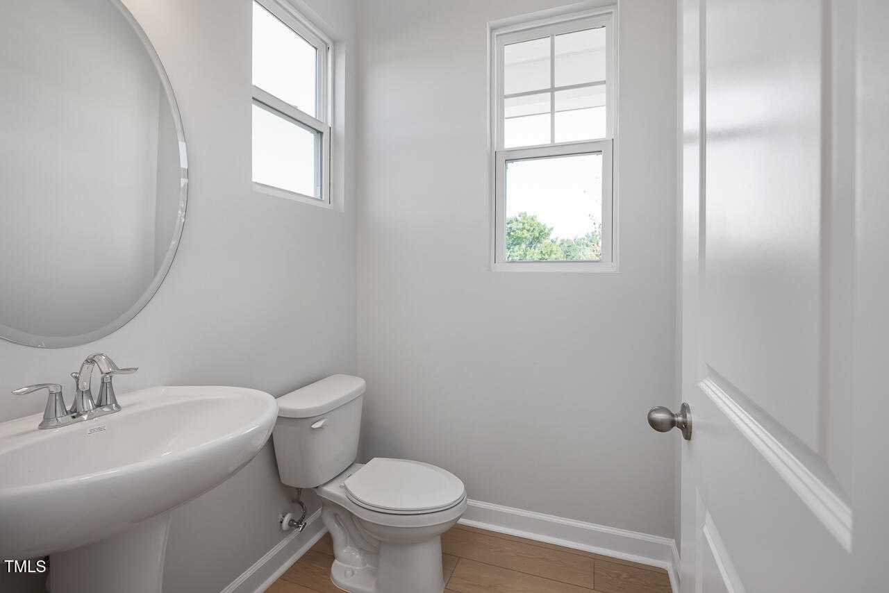 Bright powder room with round mirror, white sink, toilet, window view, and hardwood floor in Davidson Homes The Graham, Fuquay-Varina, NC
