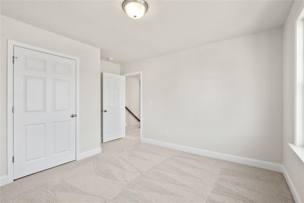 Bright empty bedroom with white walls, beige carpet, closet door, and hallway access in Davidson Homes The Ash C, Hoschton, GA