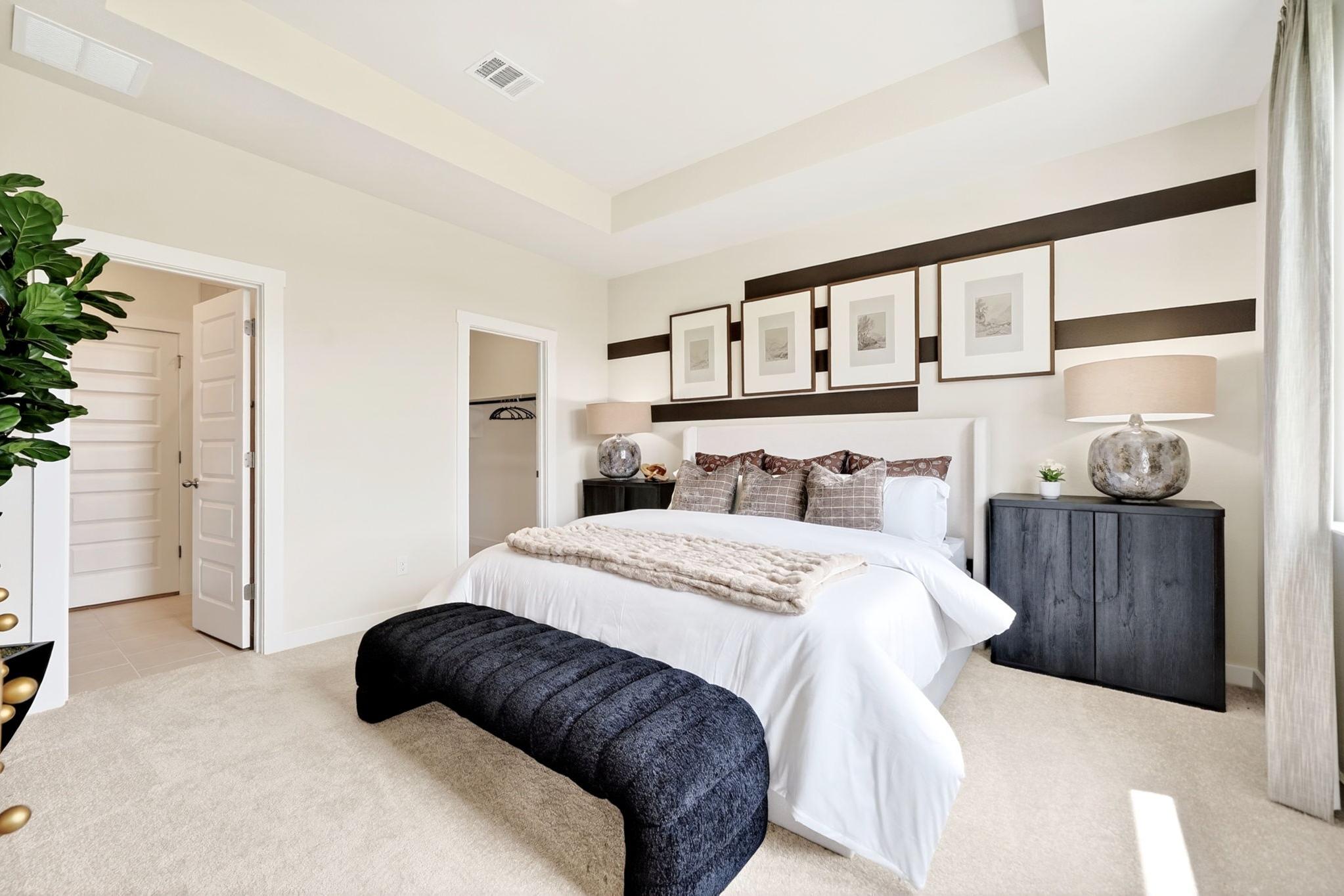 Spacious master bedroom in Heartland Texas by Davidson Homes with king bed, black artwork, plush bench, and potted fiddle leaf fig
