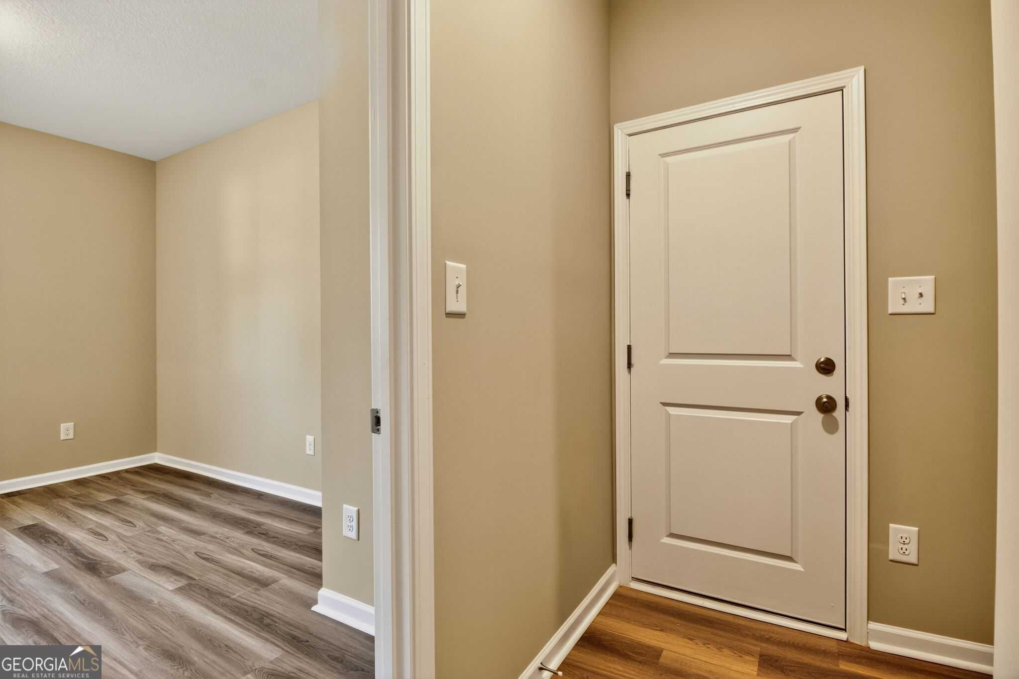 Spacious empty bedroom with beige walls, luxury vinyl plank floors, and adjacent hallway white door in Evermore Homes The Luna, Perry GA