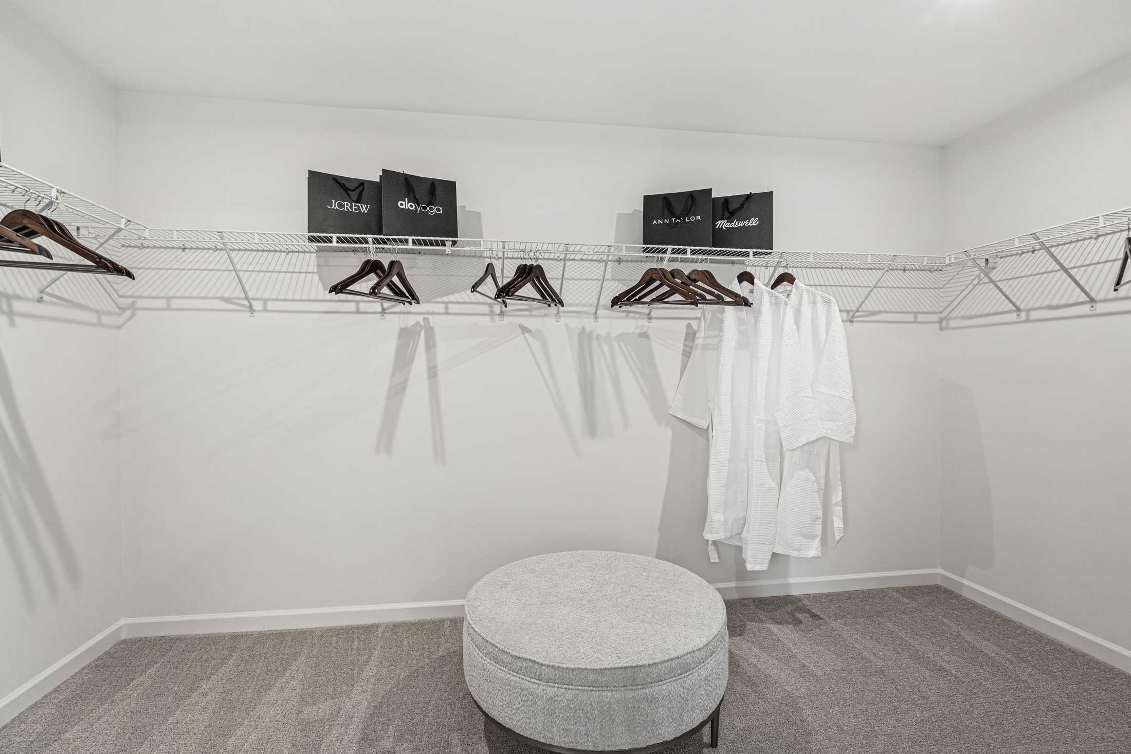 Spacious walk-in closet in The Hickory A master suite with wire shelving, hanging white robes, black storage bags, and central gray ottoman on carpet