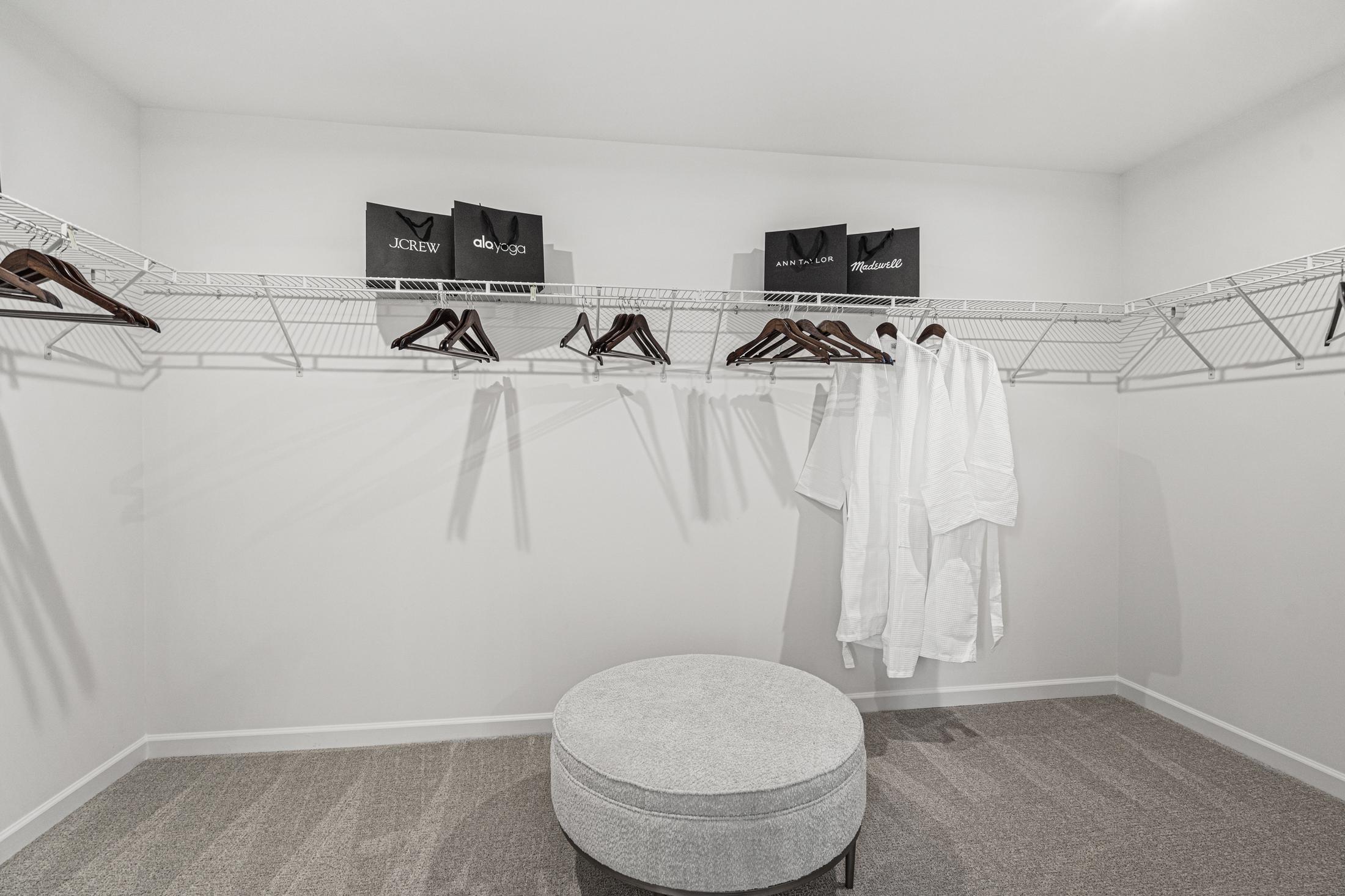 Spacious walk-in master closet in The Hickory B with wire shelving, hanging white robes, black storage bins, and gray ottoman on carpet