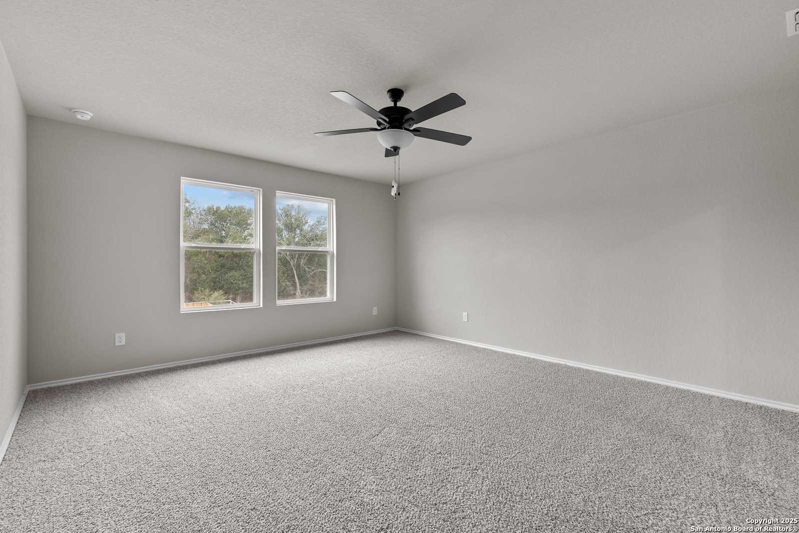 Bright bedroom with neutral gray walls, beige carpet, ceiling fan, and large windows overlooking greenery in Davidson Homes The Murray I, San Antonio