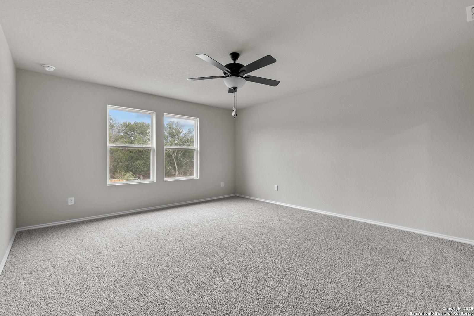 Bright bedroom with neutral gray walls, beige carpet, ceiling fan, and large windows overlooking greenery in Davidson Homes The Murray I, San Antonio