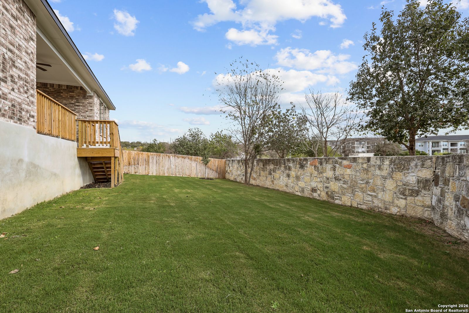 Expansive backyard with wooden deck, stairs, lush green lawn, and stone walls in Davidson Homes The Lanier H, Ladera, San Antonio