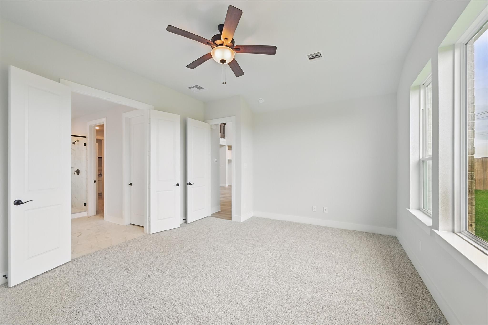 Spacious carpeted master bedroom with ceiling fan, large windows, and open en-suite bathroom doors in Davidson Homes The Victoria C, Texas City