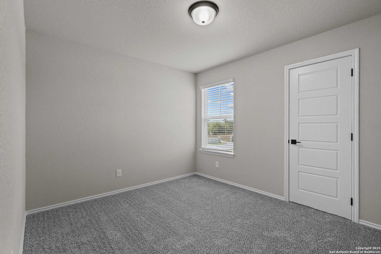 Bright secondary bedroom with light gray walls, gray carpet, white door, and window blinds in Davidson Homes The Charlotte B, San Antonio