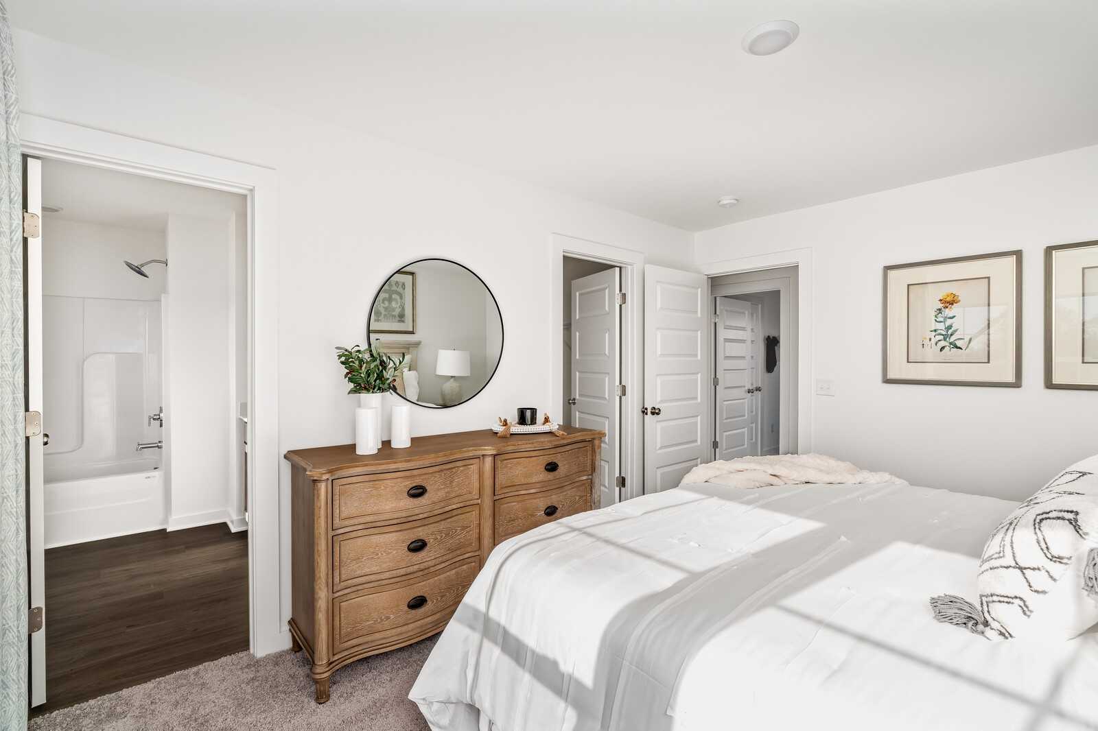 Cozy master bedroom with oak dresser, white duvet bed, round mirror, botanical art, and en-suite bath with shower tub in Davidson Homes Cumberland A, Gallatin TN