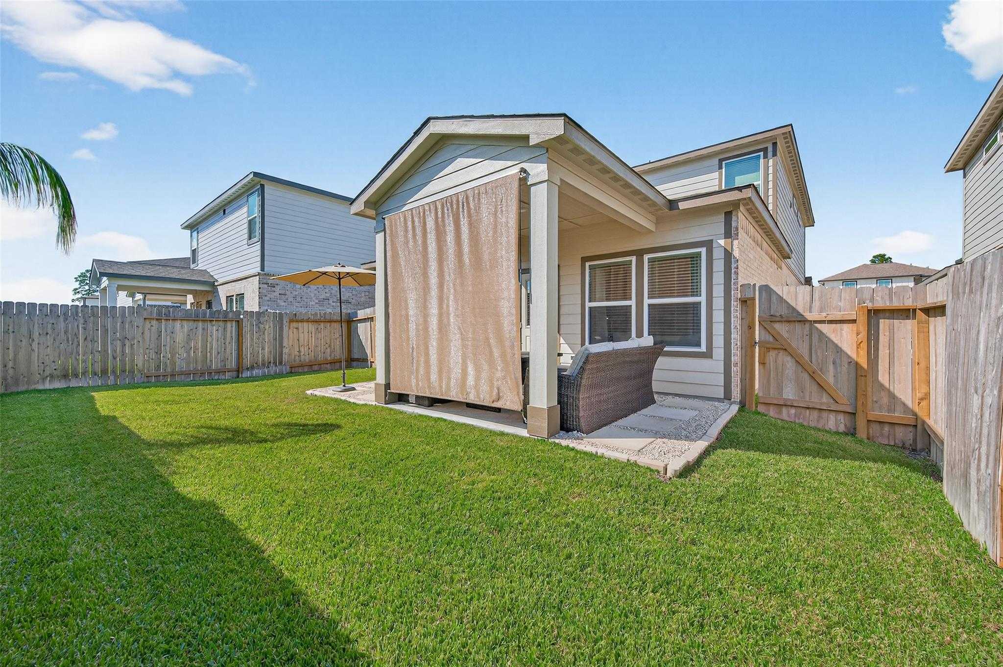 Covered patio with drop-down beige shades, cushioned outdoor seating, lush green lawn, and privacy fence in Lakes at Black Oak home, Magnolia, Texas