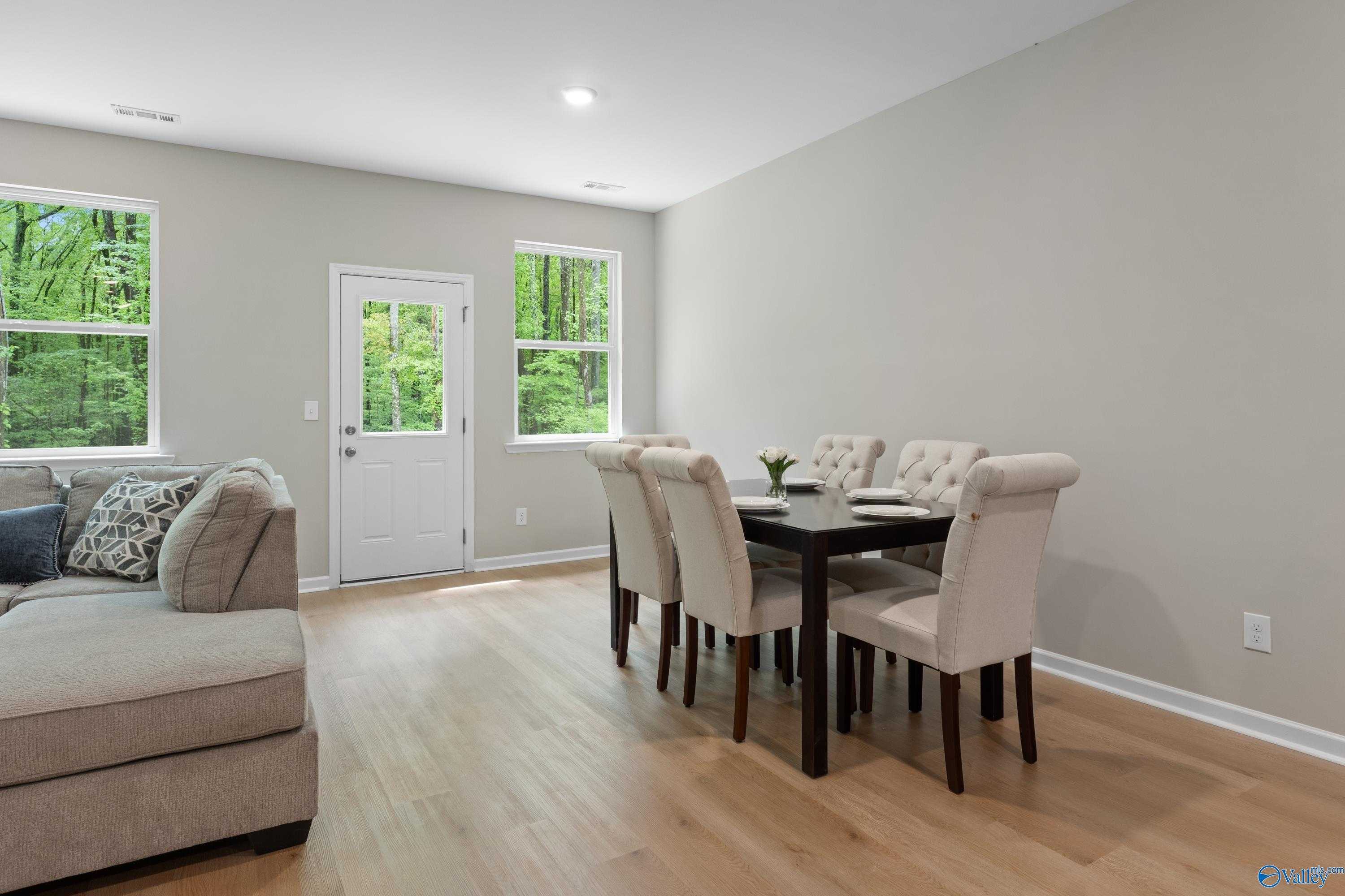 Open living-dining area with L-shaped beige sofa, dark wood table, upholstered chairs, French doors to wooded view in Davidson Homes The Luna, Hazel Green, AL