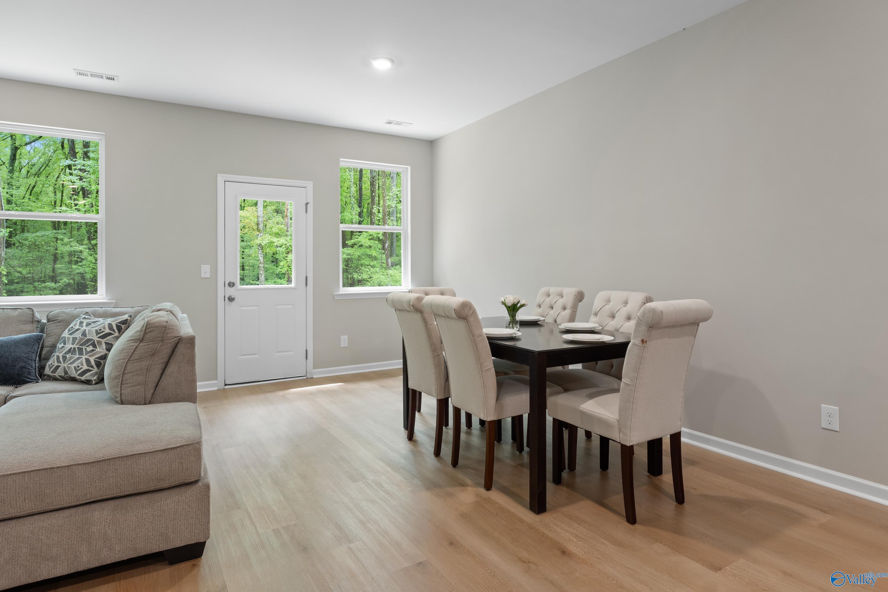 Open-concept living-dining area with L-shaped beige sofa, wood table, tufted chairs, large windows overlooking trees in The Luna home, Hazel Green, AL