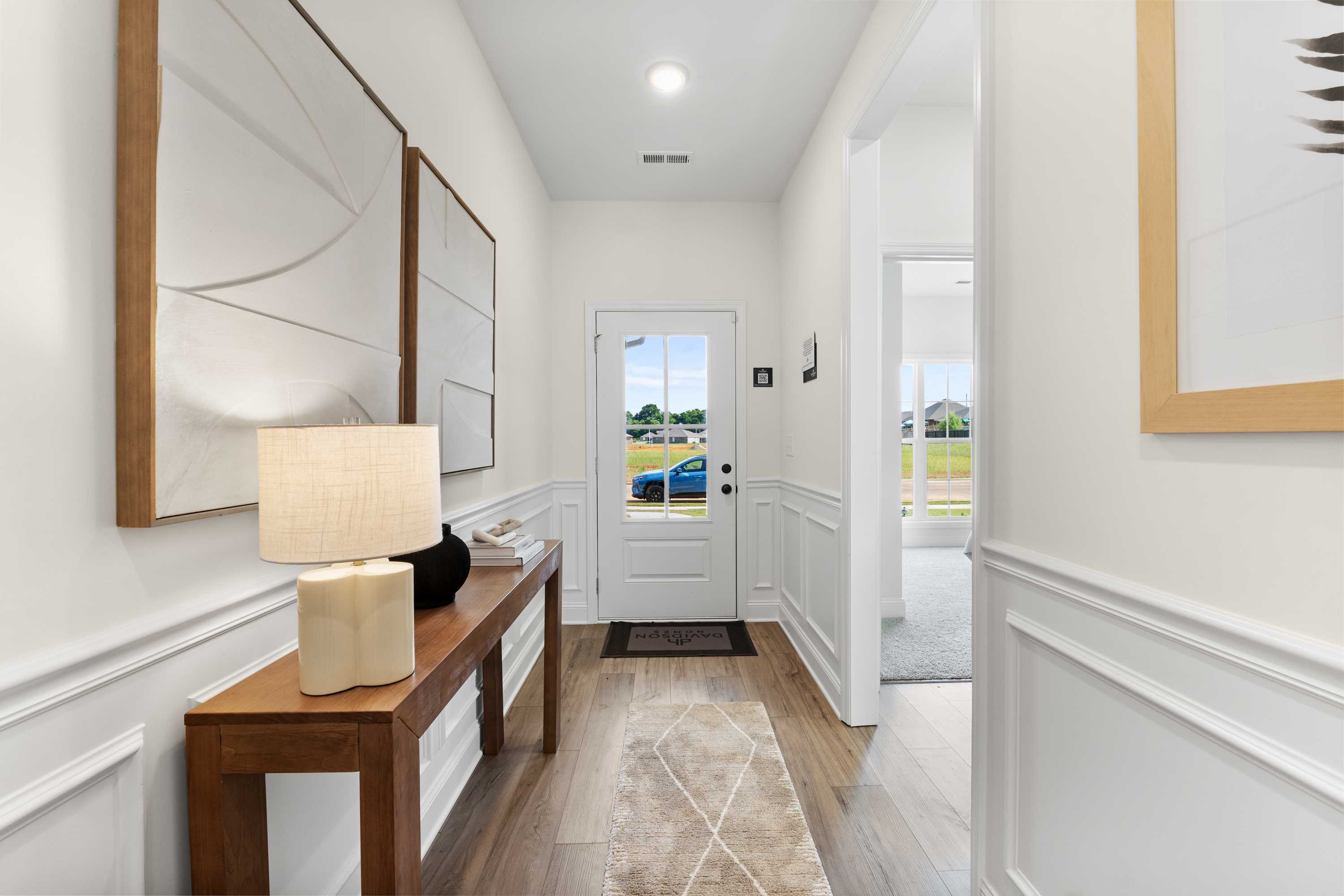Bright entry hallway in Lynn Meadows home, Meridianville Alabama with white wainscoting, wooden console table, lamp, abstract art, and front door view