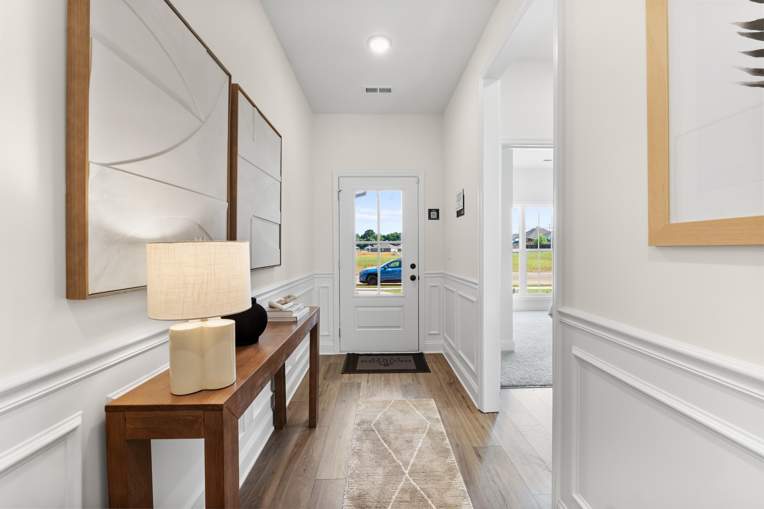 Bright entry hallway in Lynn Meadows home, Meridianville Alabama with white wainscoting, wooden console table, lamp, abstract art, and front door view