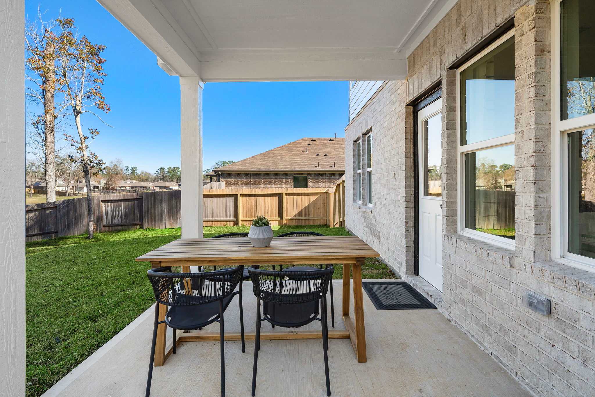 Covered patio with wooden dining table and wicker chairs at Lakes at Black Oak in Magnolia Texas featuring brick home exterior and lush green lawn