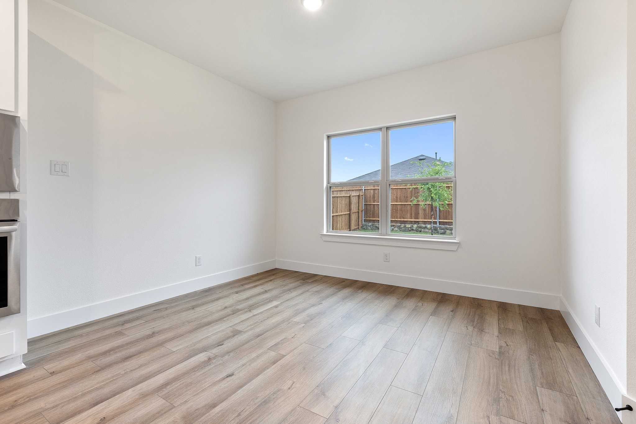 Bright empty dining area with light hardwood floors, white cabinets, stainless oven, and large windows overlooking fenced backyard in 4-bedroom Josephine Texas home