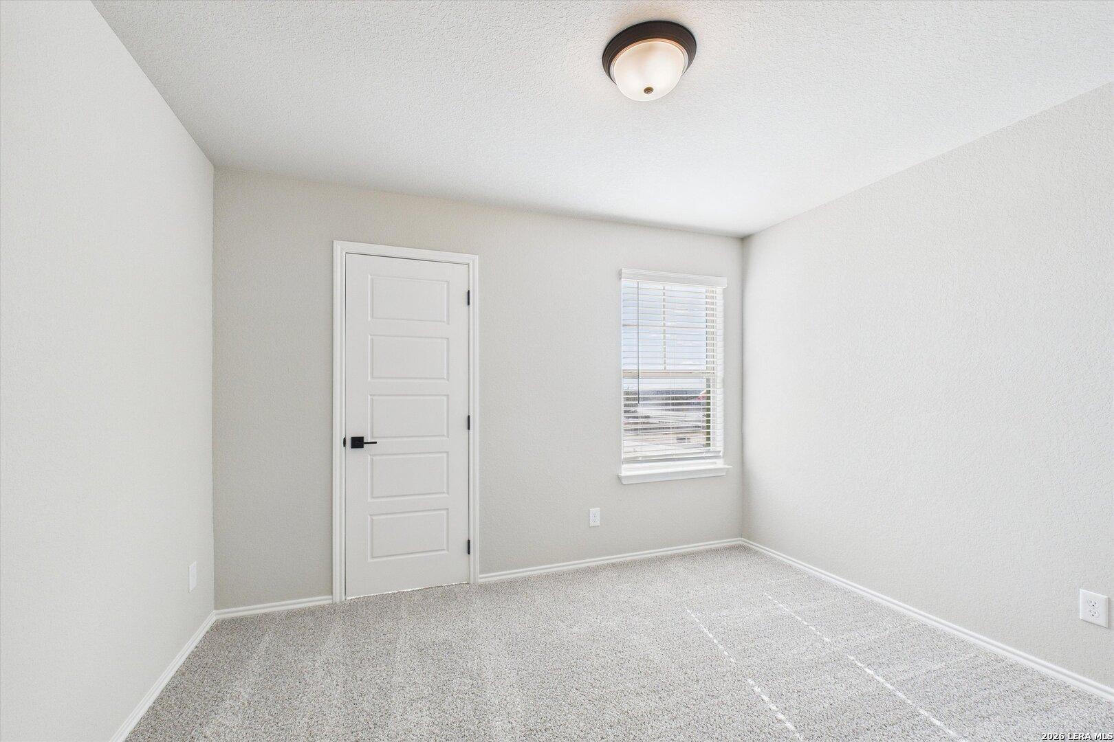 Bright empty bedroom with neutral gray walls, white door, window blinds, and plush carpet in Davidson Homes The Charlotte A, San Antonio