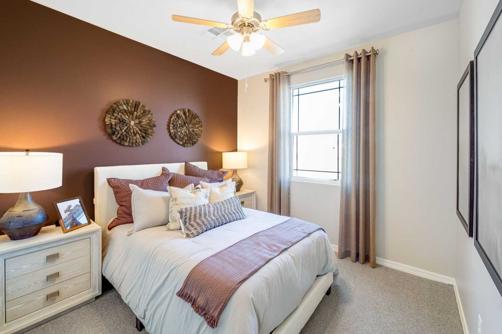Cozy master bedroom in The Savannah C featuring plush bed, brown accent wall with woven art, ceiling fan, and window curtains