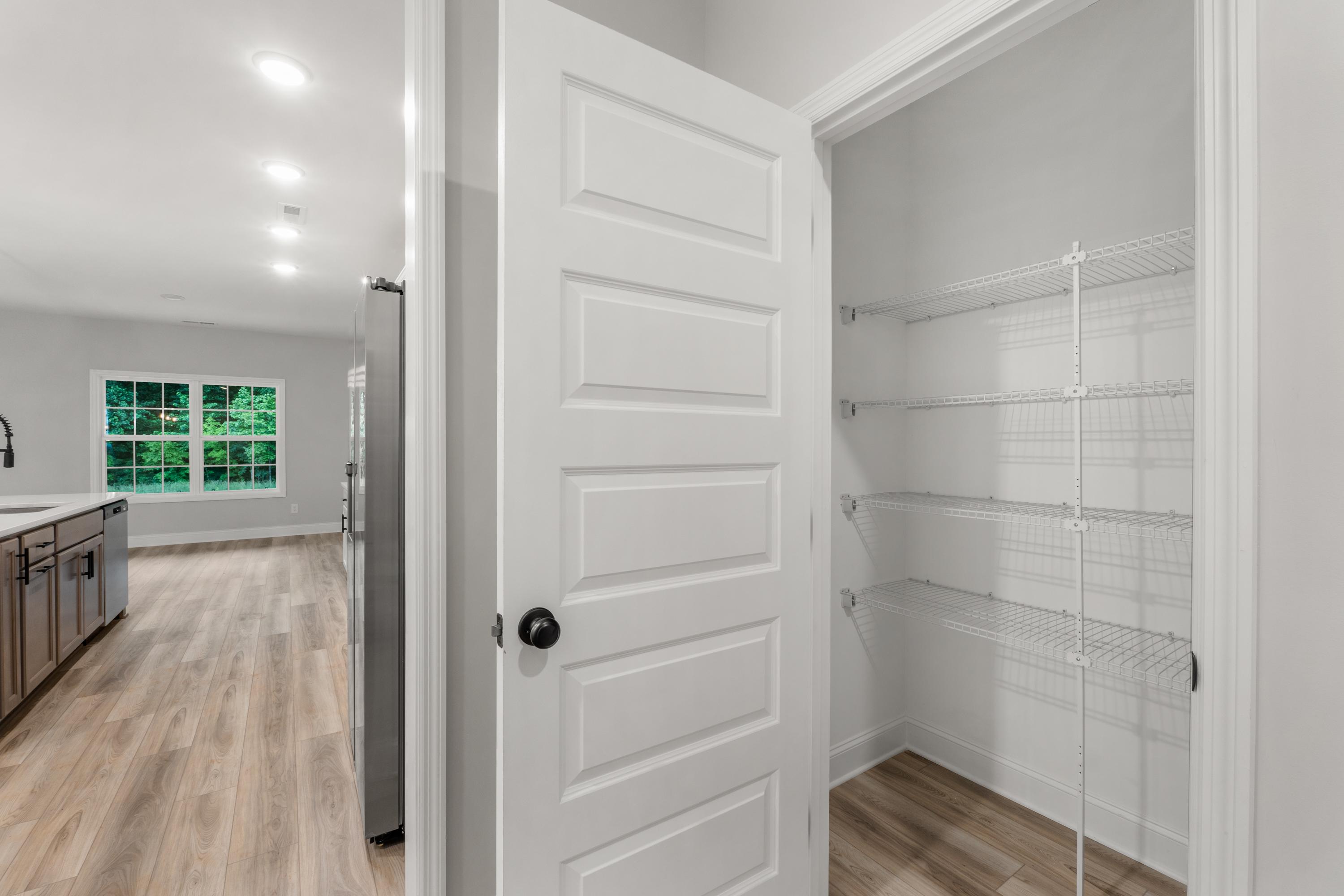 Spacious walk-in pantry in The Oxford home with open white door, wire shelving, adjacent modern kitchen, stainless fridge, and hardwood floors
