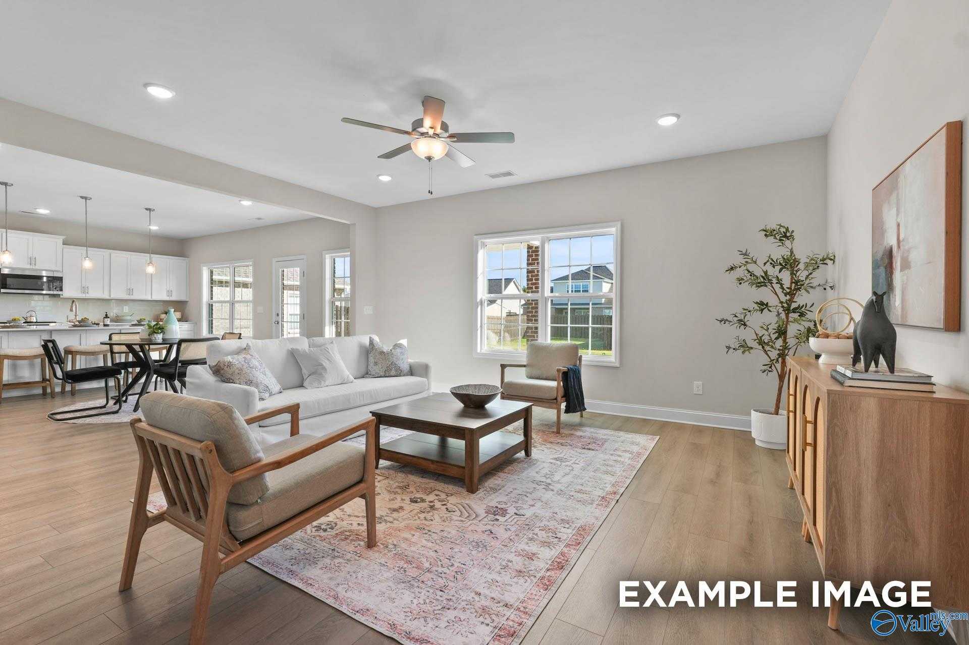 Open-concept living room with ceiling fan, large windows, and adjacent white kitchen in 5-bedroom Evermore Homes The Madison D, Madison, Alabama