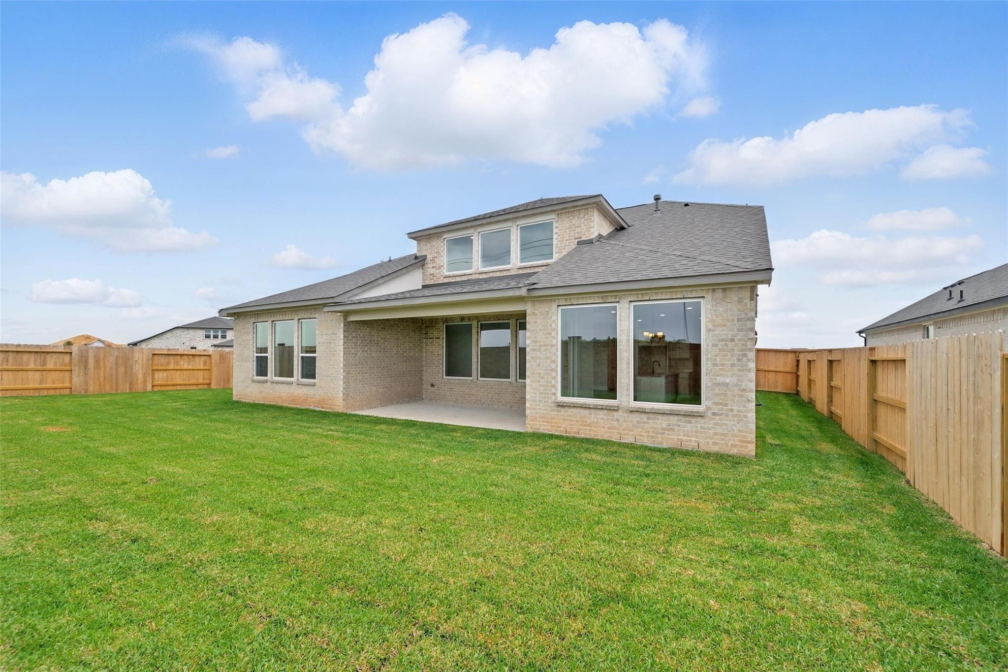 Back view of two-story tan brick home with covered patio, large windows, and expansive green backyard in Davidson Homes The Victoria C, Lago Mar, Texas City