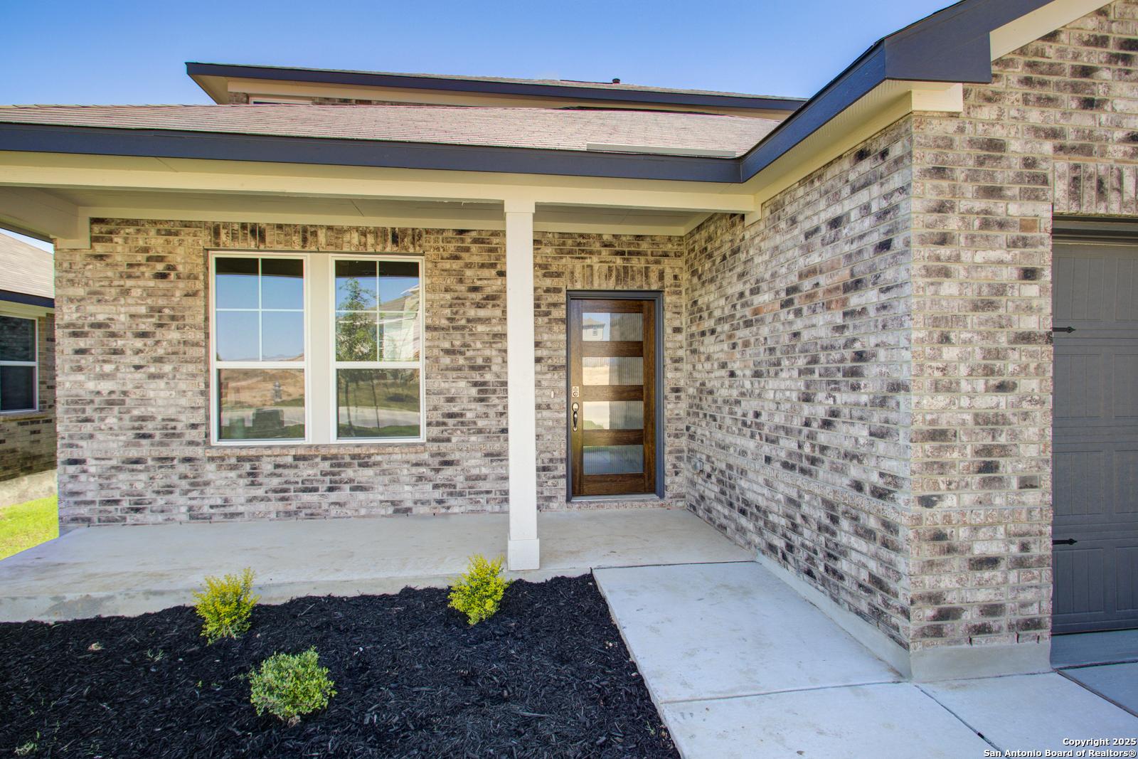 Modern beige brick home exterior with columned covered porch, frosted glass door, large windows, 2-car garage, and landscaped yard in Comanche Ridge, San Antonio