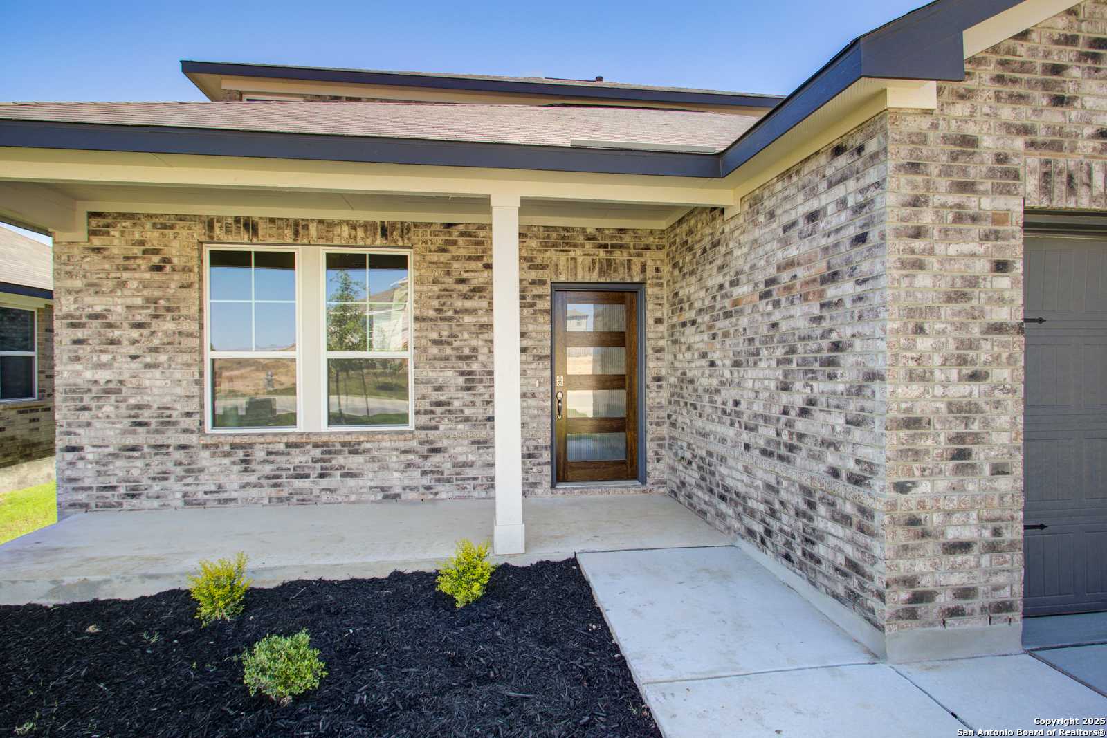 Modern beige brick home exterior with columned covered porch, frosted glass door, large windows, 2-car garage, and landscaped yard in Comanche Ridge, San Antonio