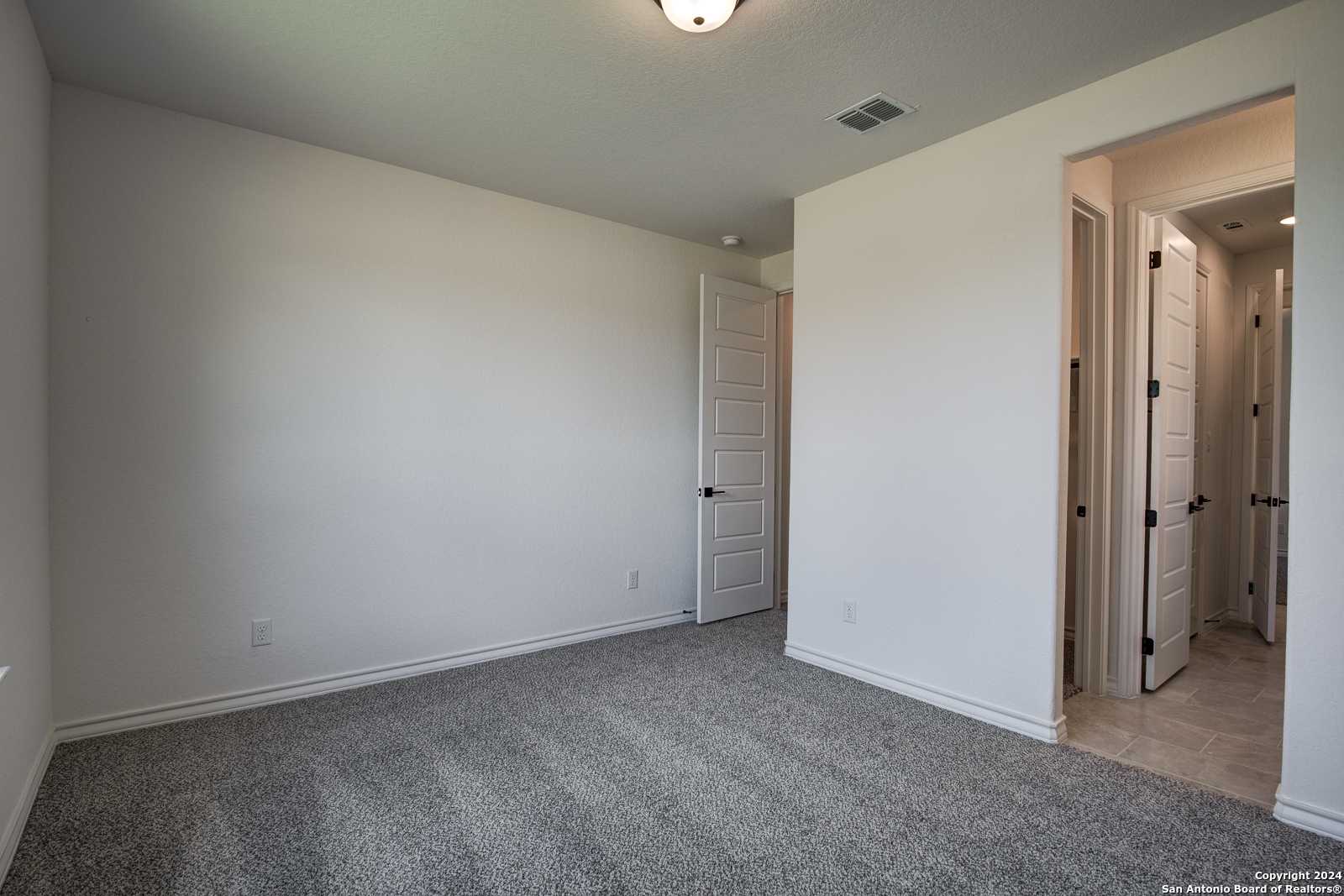 Spacious empty bedroom with gray carpet, white walls, and adjacent tiled bathroom in The Garner B 4-bedroom home, Castroville, Texas