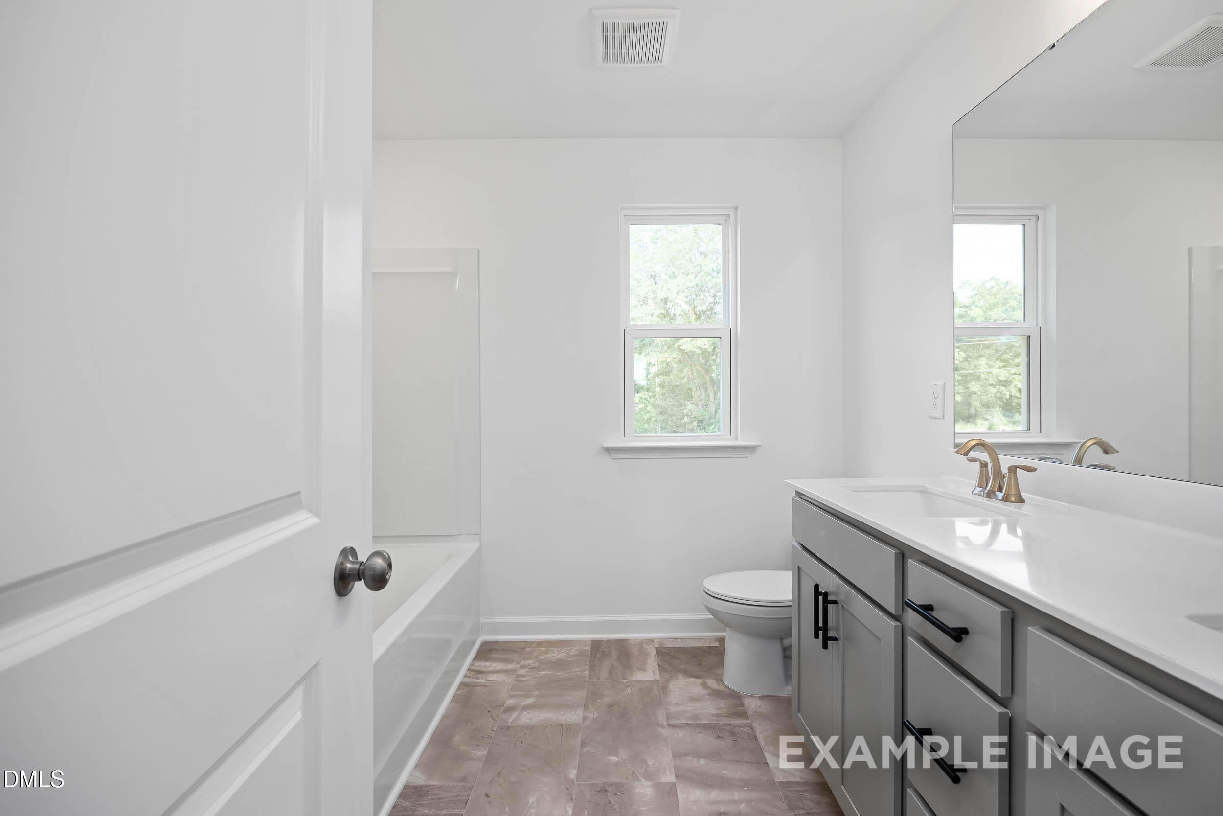 Modern bathroom with white soaking tub, gray vanity, gold faucet, and window in The Willow D by Davidson Homes, Zebulon, NC