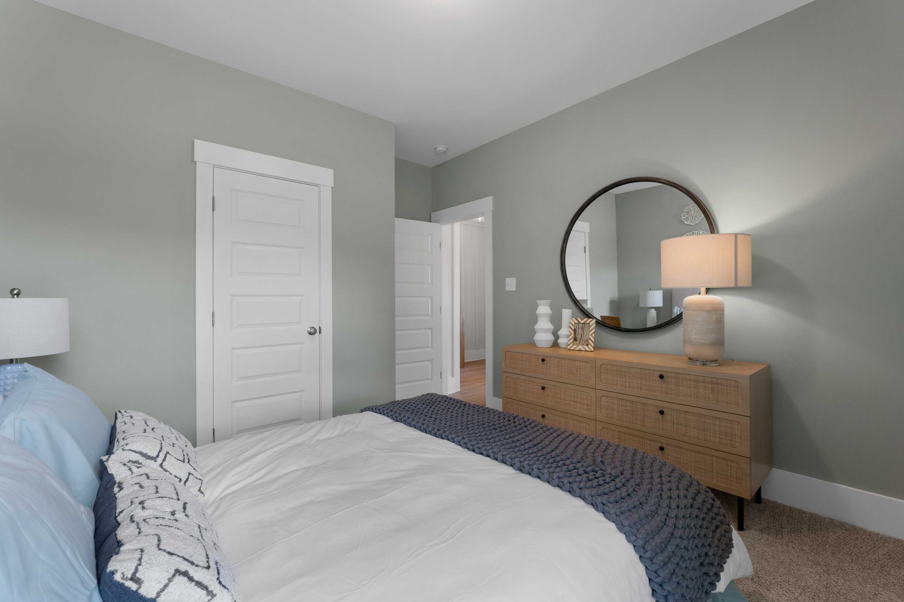 Spacious bedroom interior at Hollon Meadow in Decatur Alabama featuring light gray walls, king bed with white bedding, wooden dresser, and round mirror