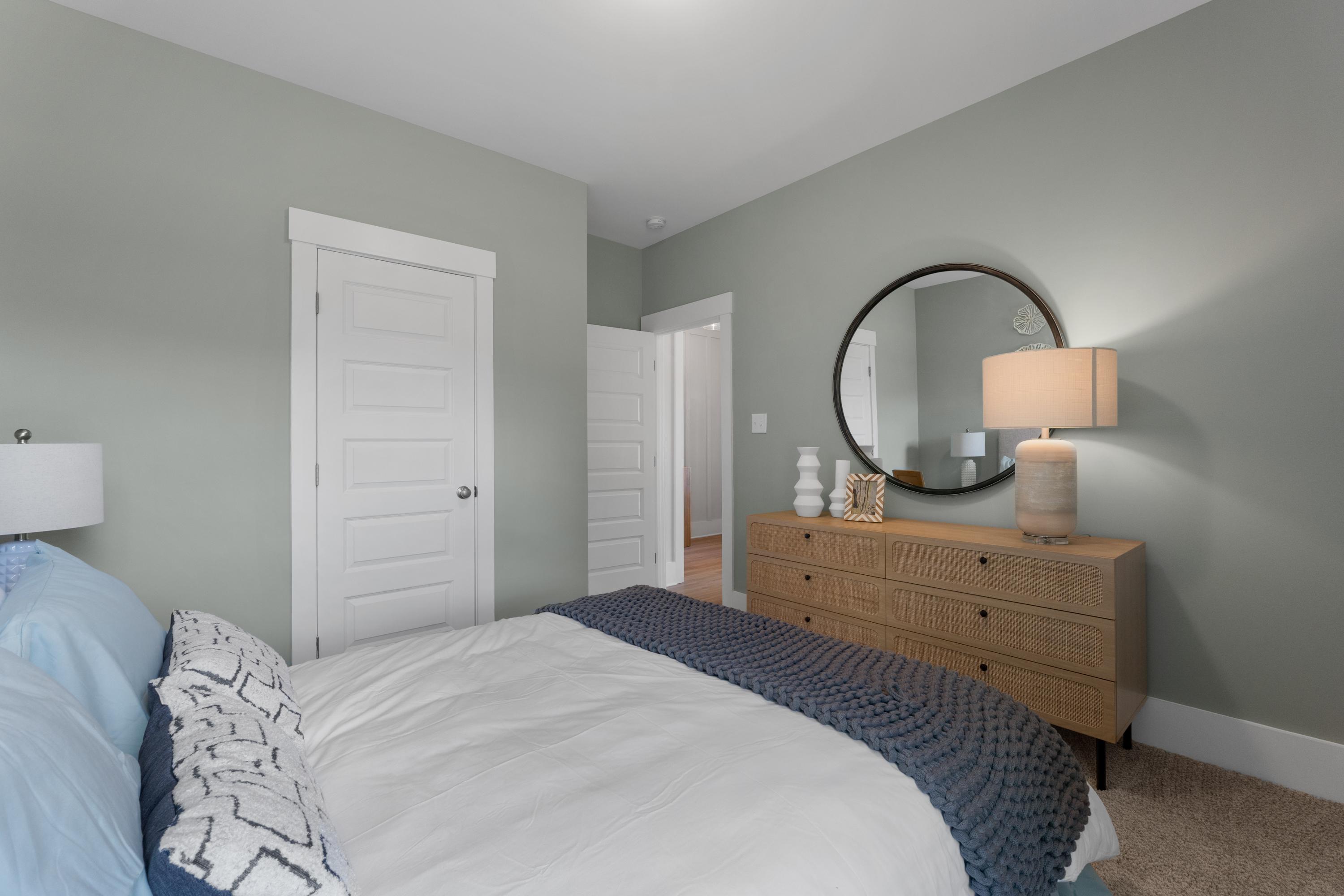 Spacious bedroom interior at Hollon Meadow in Decatur Alabama featuring light gray walls, king bed with white bedding, wooden dresser, and round mirror