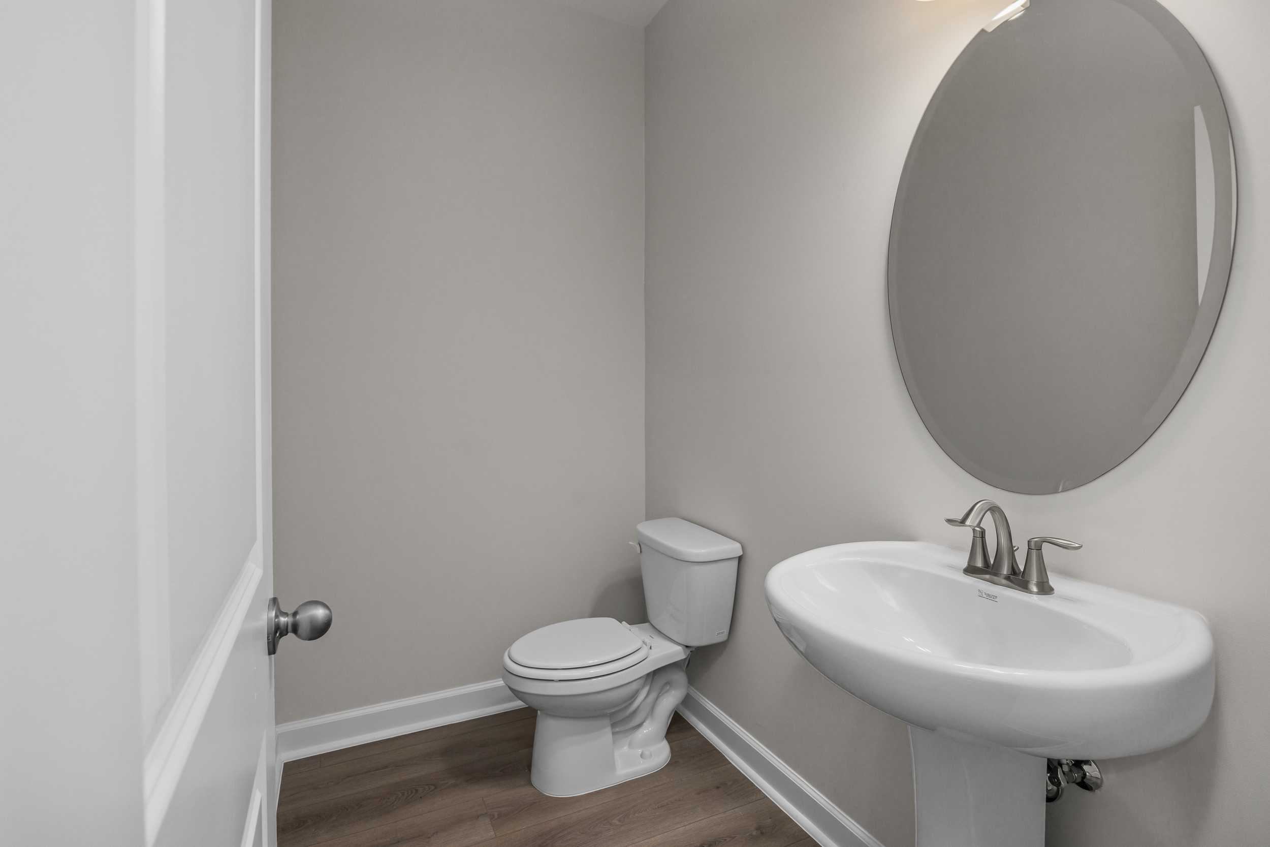 Neutral powder room in The Aspen by Davidson Homes: white pedestal sink, round mirror, toilet, gray walls, wood floors