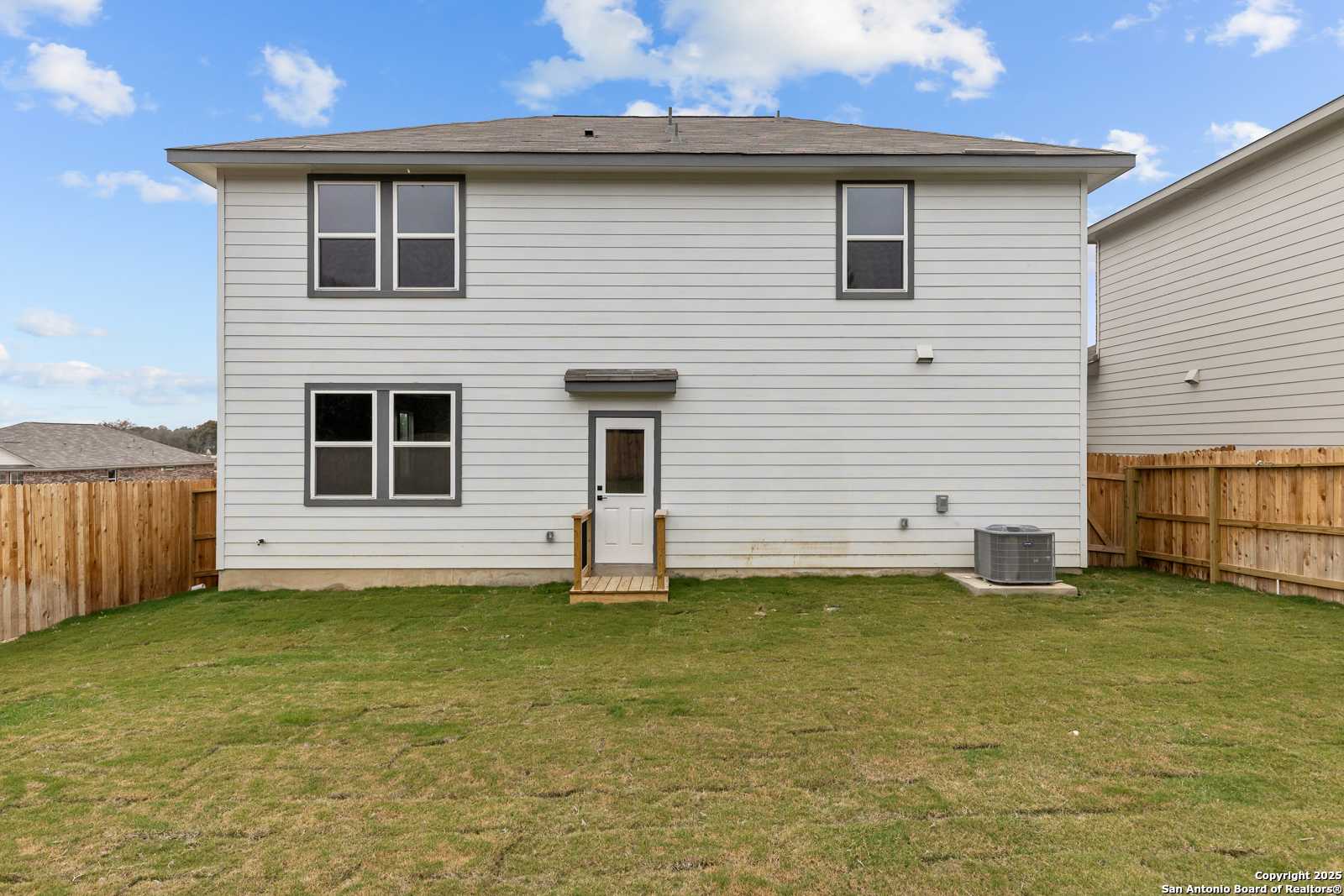Rear view of two-story white-sided home with covered patio, wooden deck, and grassy fenced backyard in Royal Crest, San Antonio, Texas