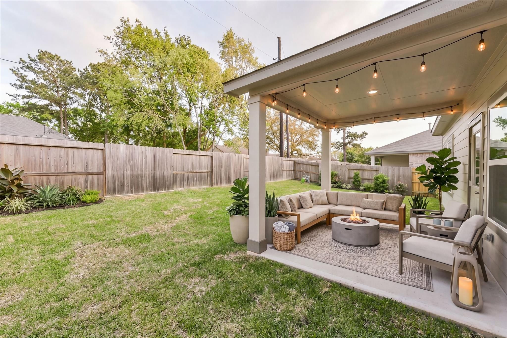 Covered back patio with L-shaped sofa, gas fire pit, string lights, and potted plants in Davidson Homes The Brazos E, Magnolia, Texas