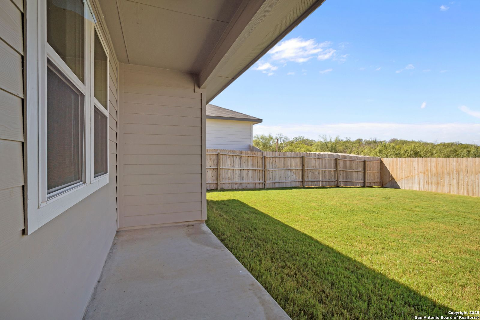 Fenced backyard with lush green lawn and covered side patio beside beige single-story home in Horizon Pointe, Converse, Texas