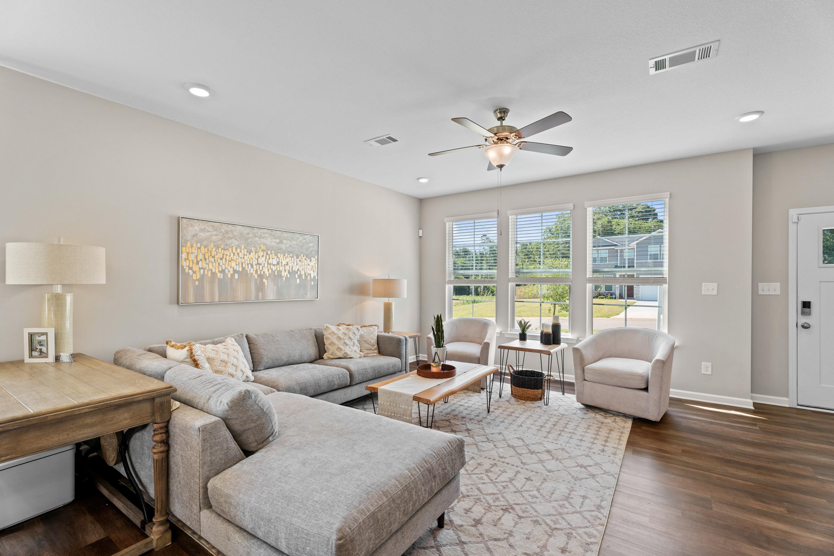 Spacious modern living room in Ivy Glen Perry GA by Evermore Homes with gray L-shaped sofa, hardwood floors, ceiling fan, large windows and abstract art