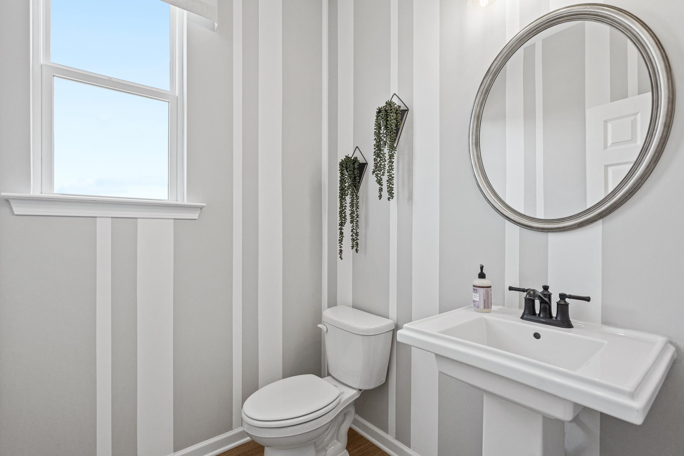 Stylish powder room in The Hickory C featuring gray striped walls, round mirror, hanging plants, pedestal sink, and toilet