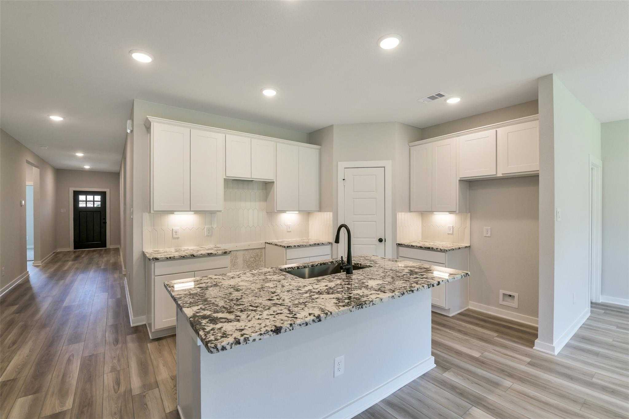 Open-concept kitchen with white cabinets, speckled granite island sink, and hardwood floors in Davidson Homes The Daphne H, Crosby Texas