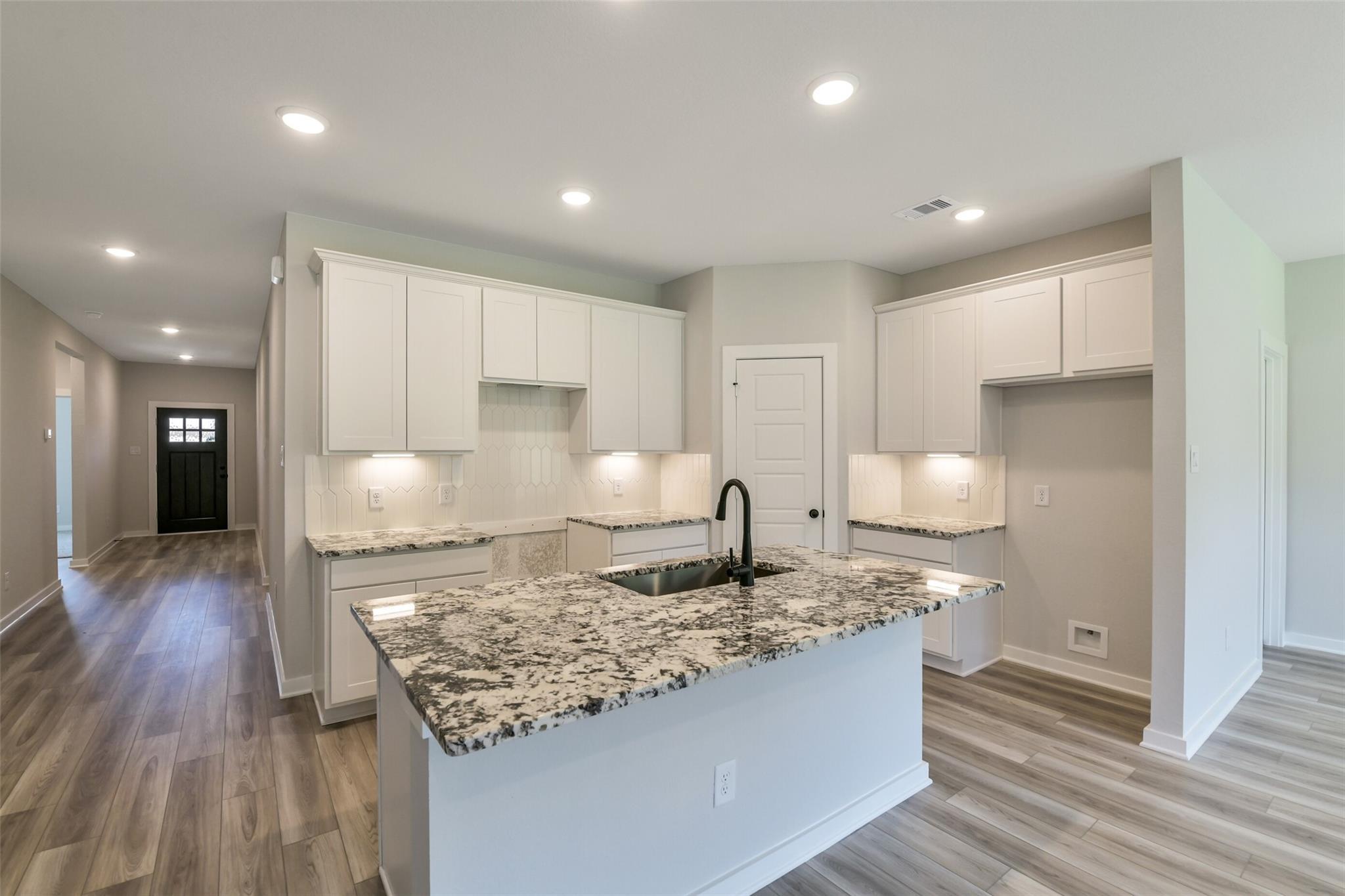 Open-concept kitchen with white cabinets, speckled granite island sink, and hardwood floors in Davidson Homes The Daphne H, Crosby Texas