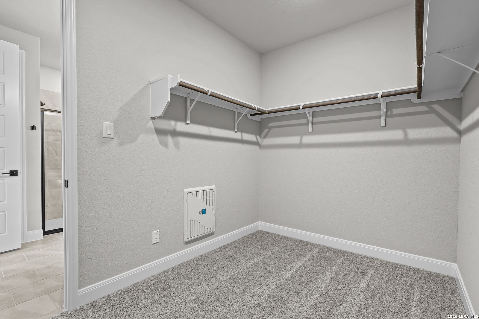 Spacious walk-in closet with wooden hanging rods, shelves, and gray carpet adjacent to en-suite bathroom in Davidson Homes The Belmont H, Castroville, Texas
