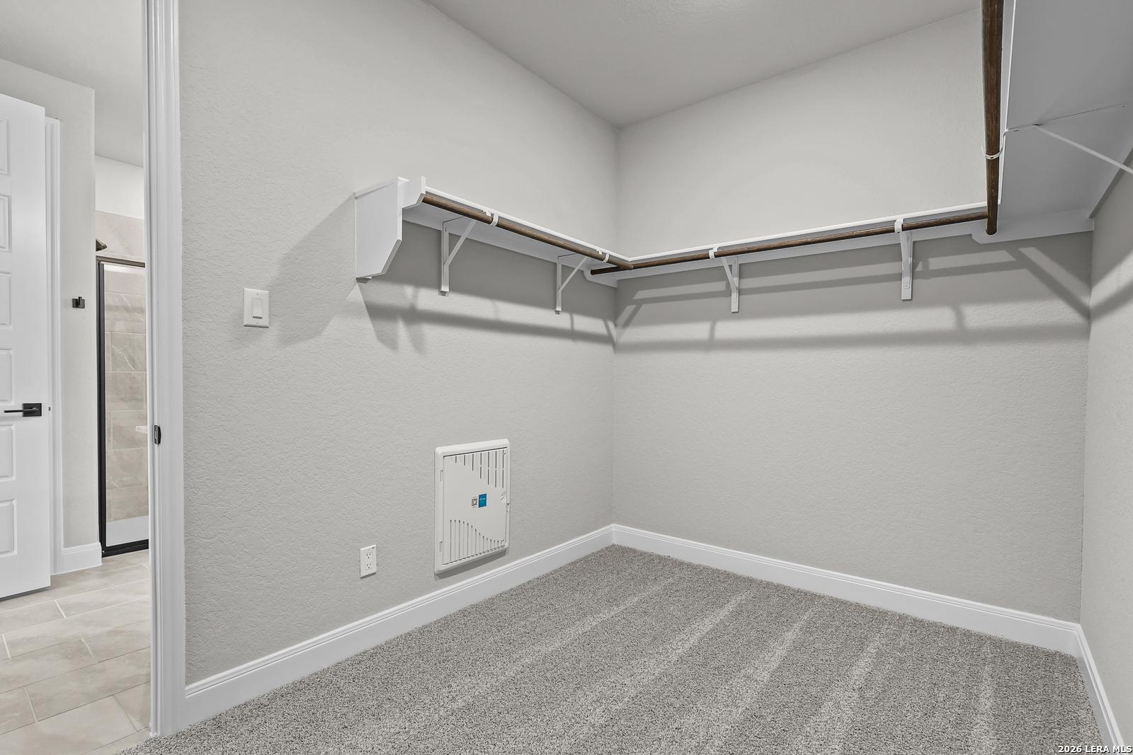 Spacious walk-in closet with wooden hanging rods, shelves, and gray carpet adjacent to en-suite bathroom in Davidson Homes The Belmont H, Castroville, Texas