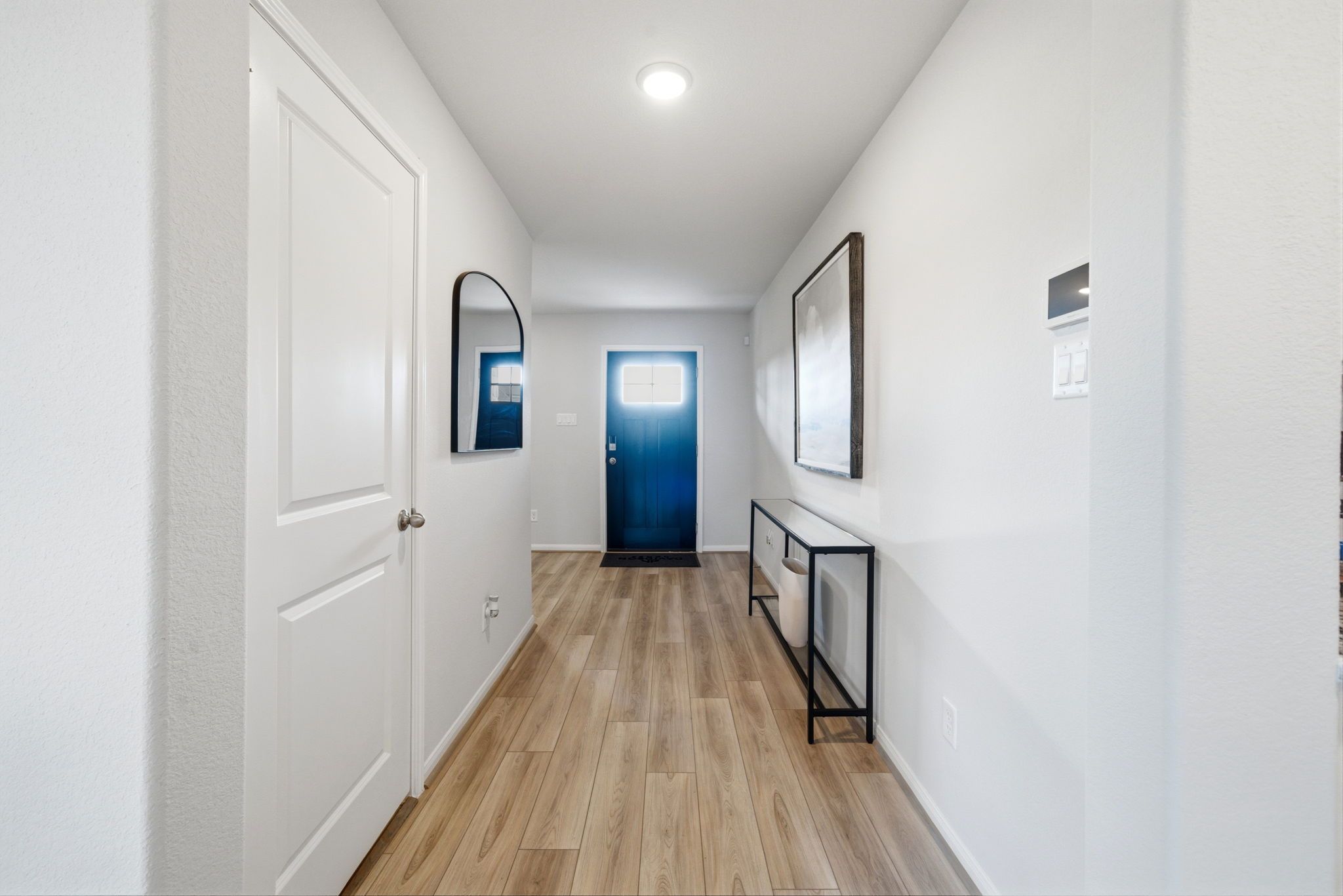 Bright hallway at Sunterra in Katy Texas featuring white walls, light hardwood floors, round mirror, framed art, black console, blue door