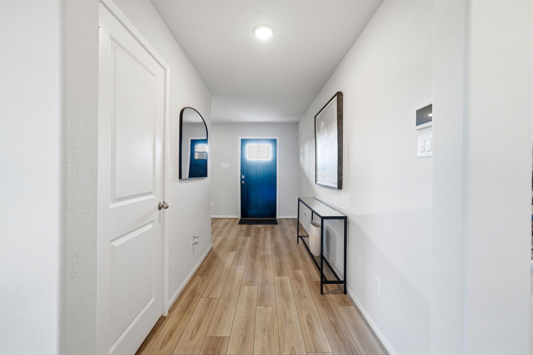 Bright hallway at Sunterra in Katy Texas featuring white walls, light hardwood floors, round mirror, framed art, black console, blue door