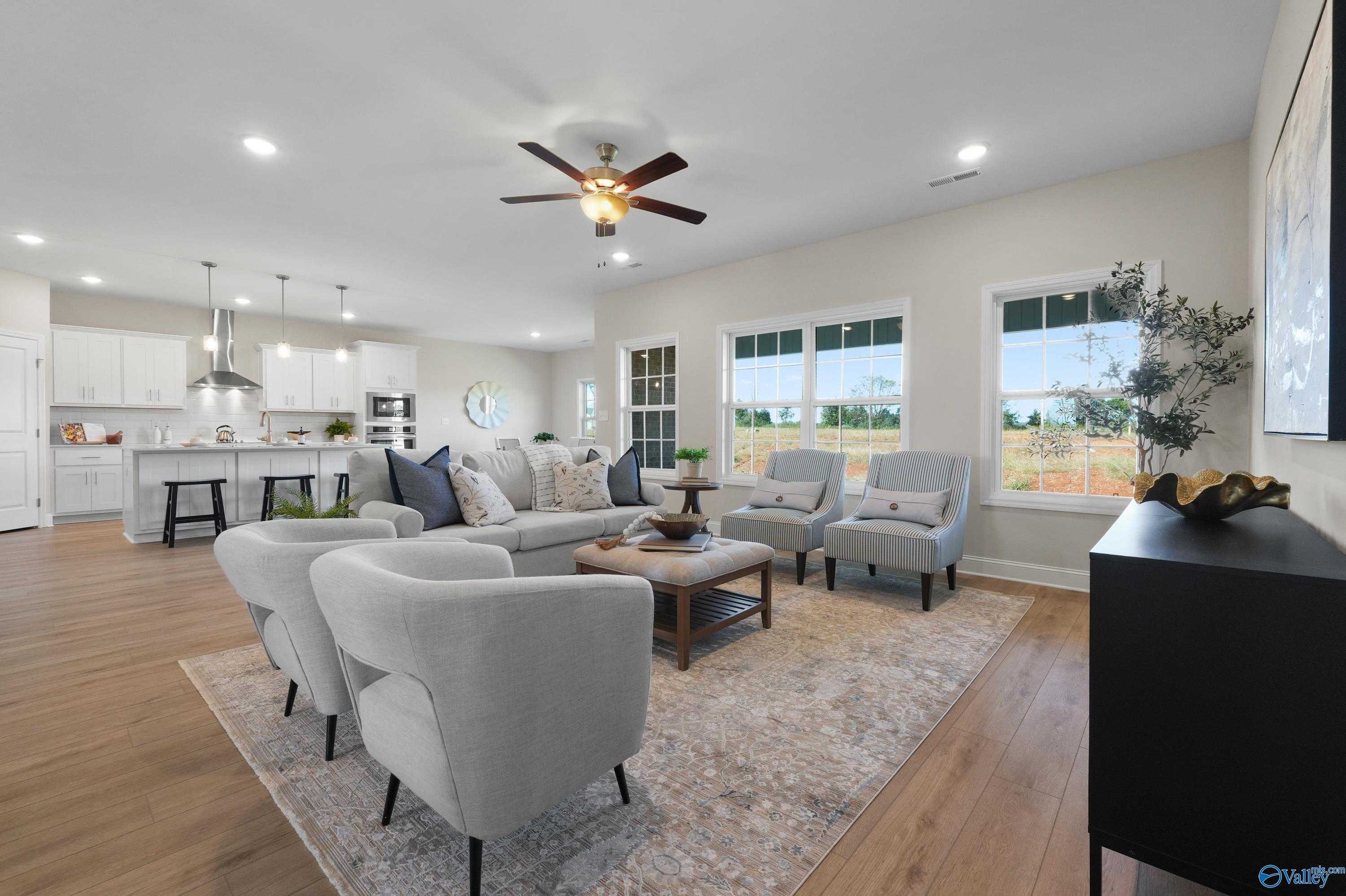 Open-concept living room with gray sectional sofa, ceiling fan, large windows, and white kitchen in Davidson Homes The Rockford B, Toney, Alabama