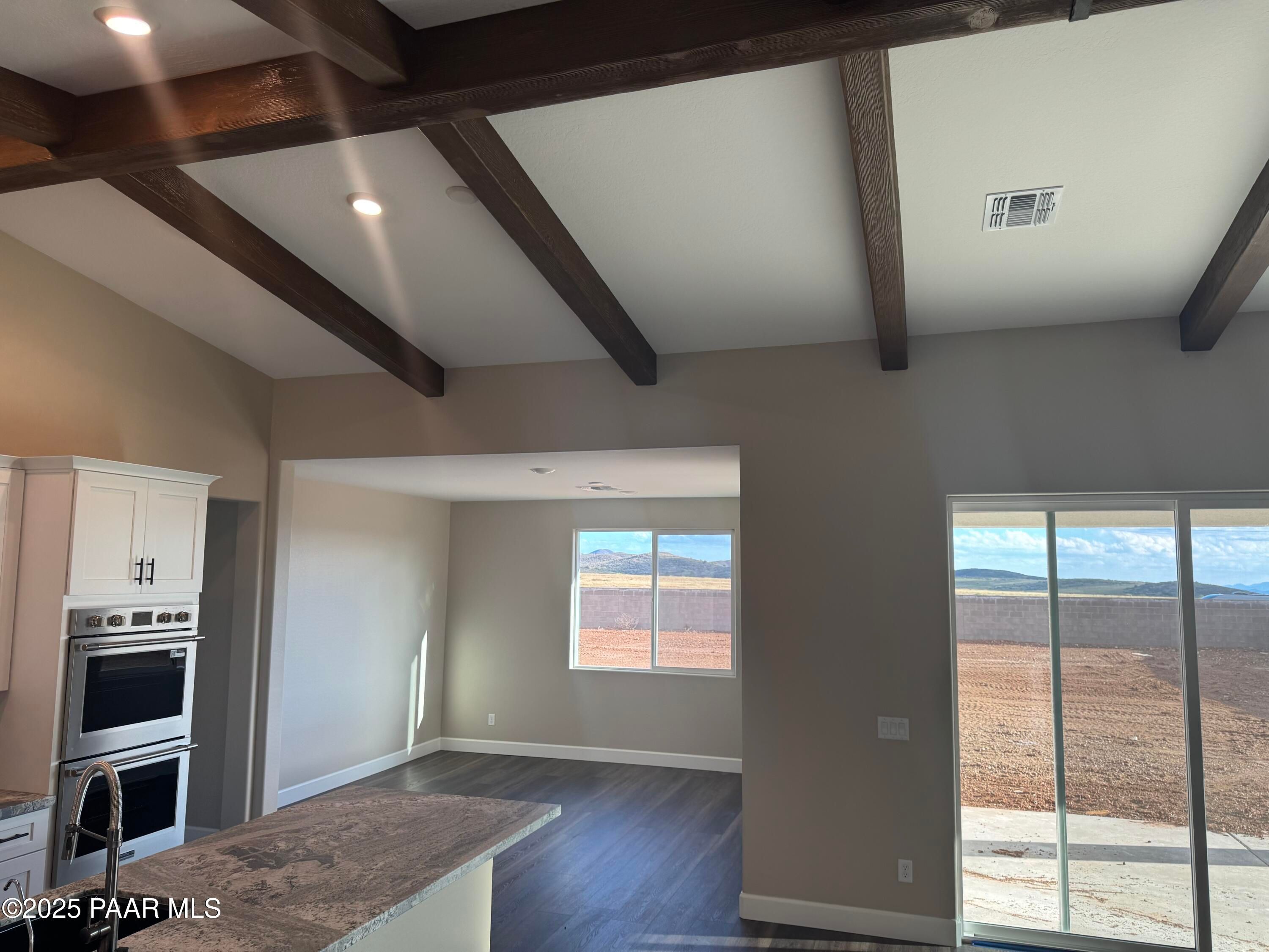 Open-concept great room with exposed wood beams, white kitchen cabinets, granite island, and desert view via sliding doors in Evermore Homes The Sunrise A, Prescott Valley, AZ