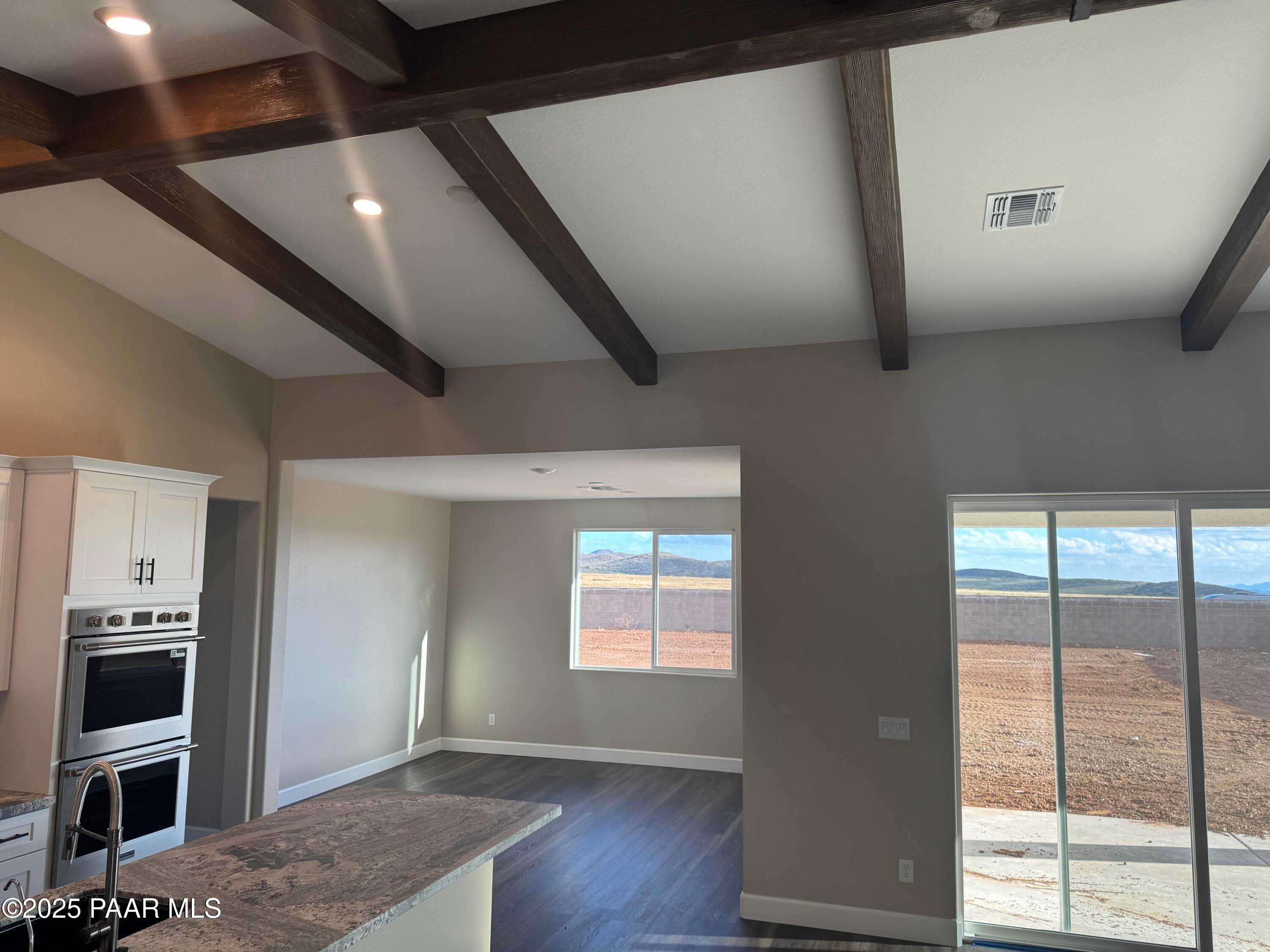 Open-concept great room with exposed wood beams, white kitchen cabinets, granite island, and desert view via sliding doors in Evermore Homes The Sunrise A, Prescott Valley, AZ