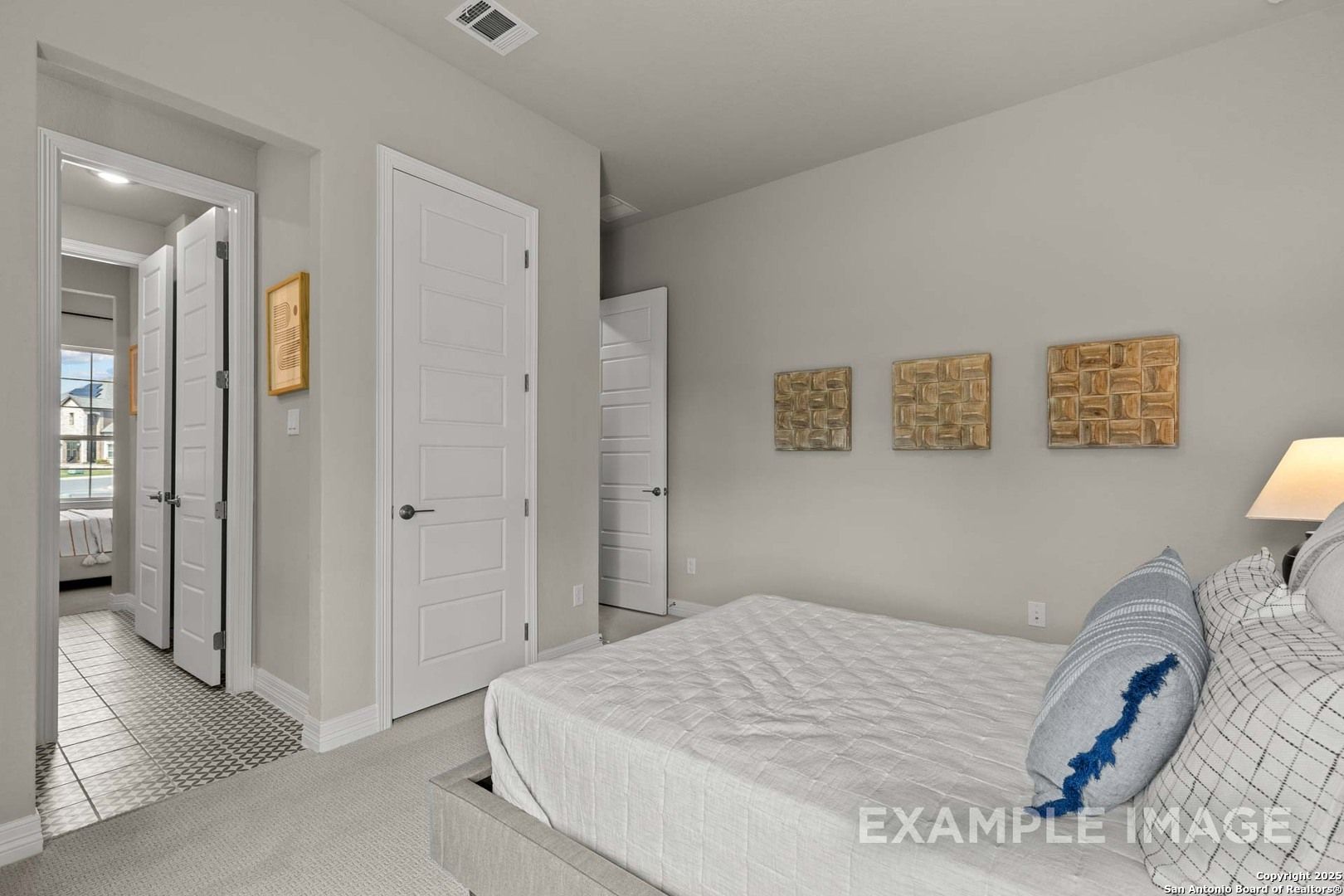 Light gray bedroom with platform bed, blue pillows, wooden art, and open bath door in Davidson Homes The Summerlin B, Castroville Texas