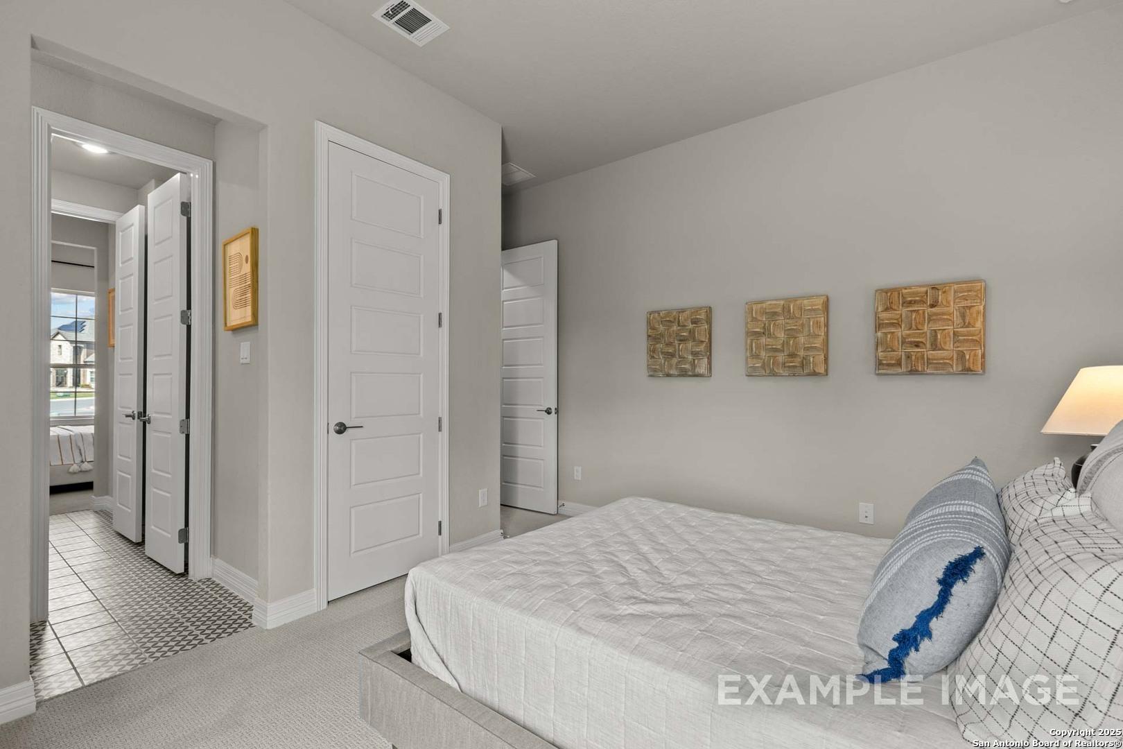 Light gray bedroom with platform bed, blue pillows, wooden art, and open bath door in Davidson Homes The Summerlin B, Castroville Texas